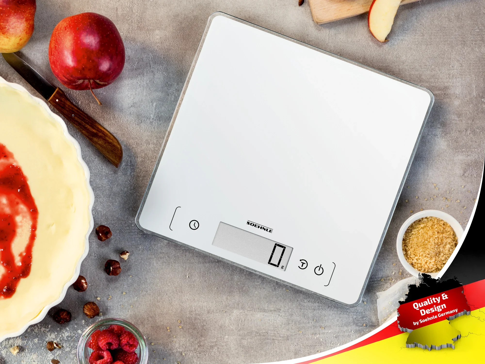 Eine digitale Küchenwaage von Soehnle liegt auf einer Arbeitsfläche, umgeben von Äpfeln, Nüssen und einer Schüssel mit Teig. Die Waage hat ein modernes, weißes Design.