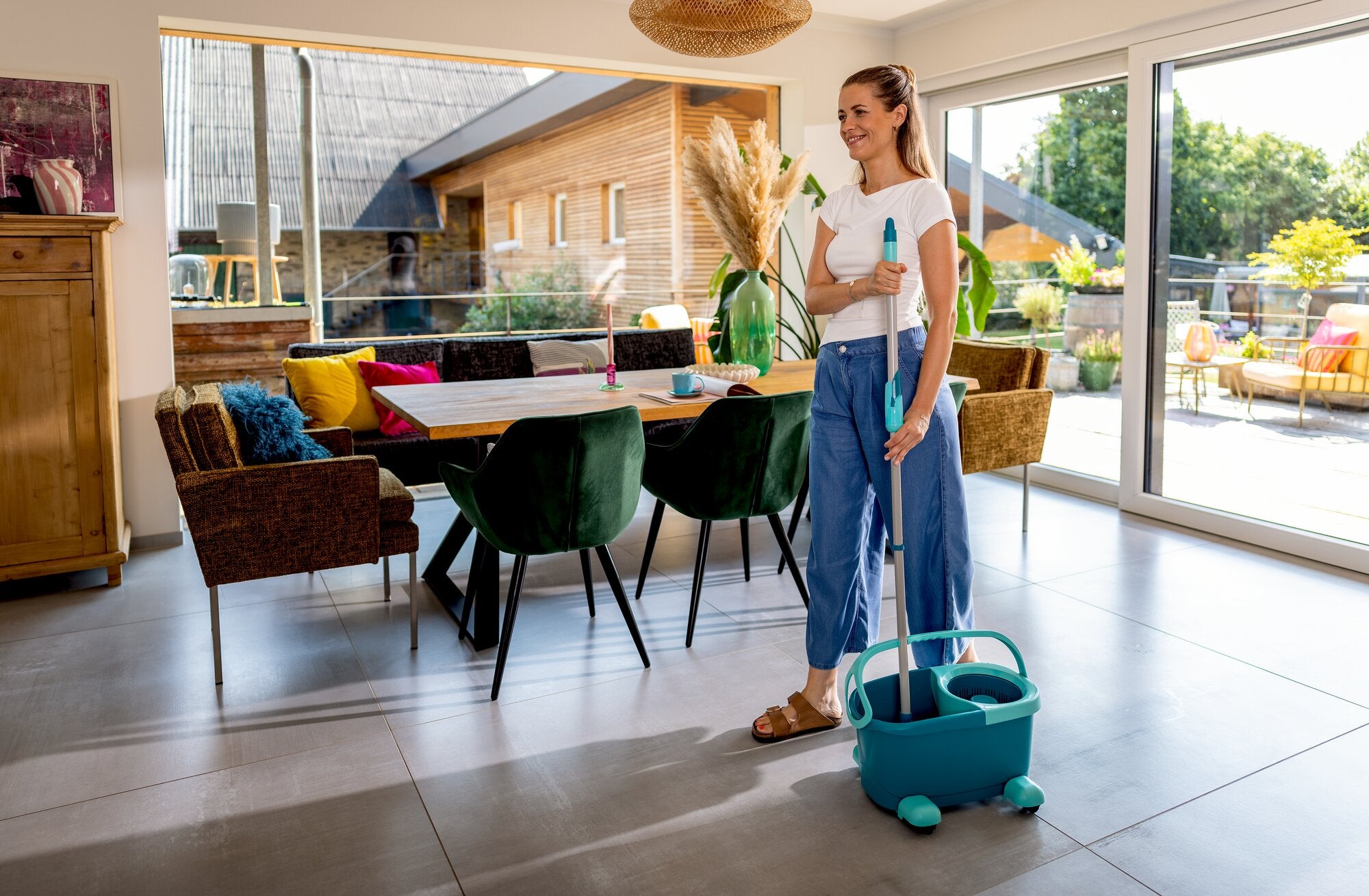 Leifheit CLEAN TWIST Disc Mop Mobile lavapavimenti mit türkisfarbenem Eimer auf Rollen im hellen Wohnraum; eine Frau wischt entspannt den Fliesenboden vor Esstisch und großer Fensterfront.