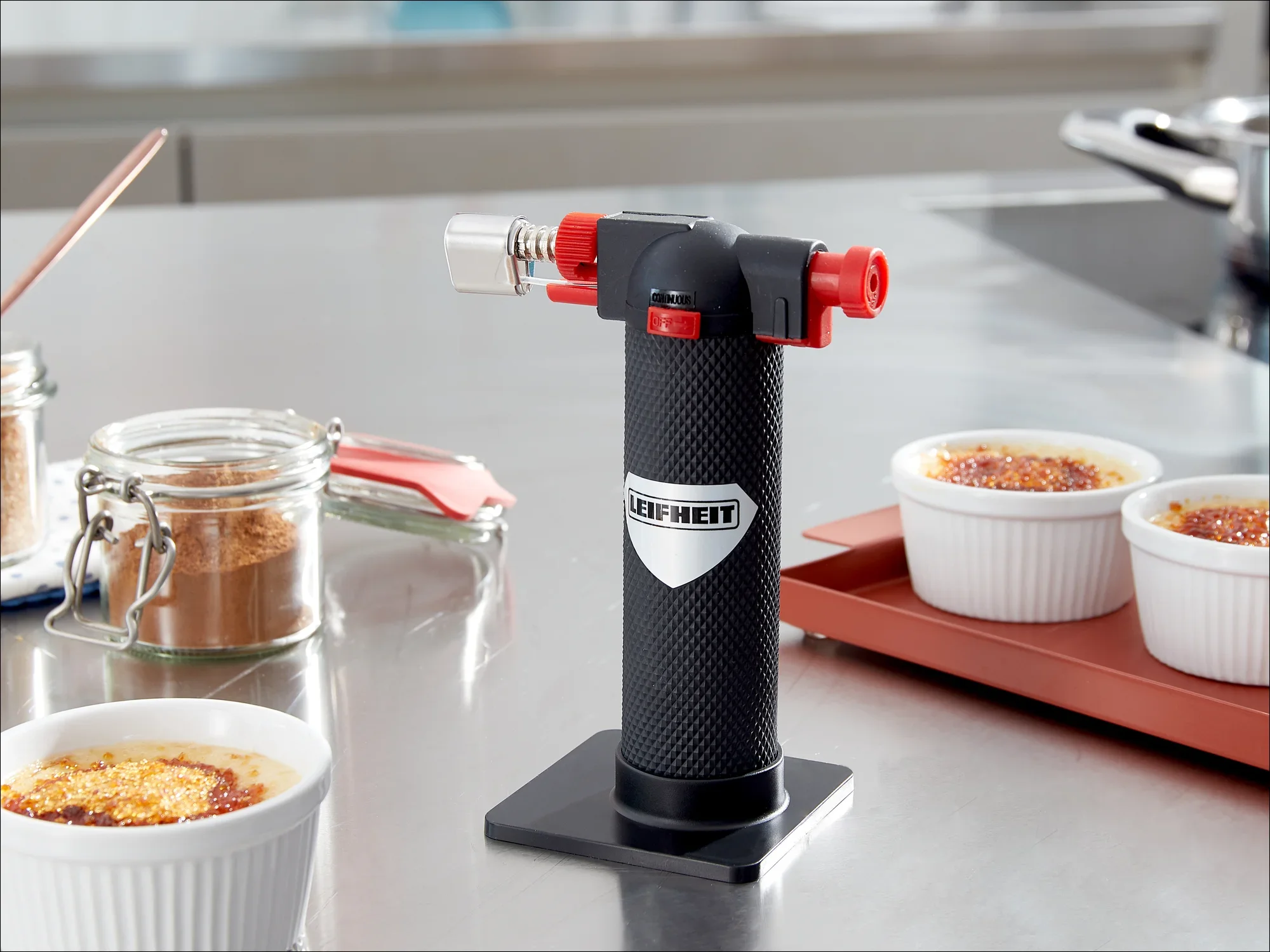 Ein Leifheit Flambierbrenner steht auf einer Küchentheke neben zwei Crème Brûlée-Schalen. Der Brenner hat ein schwarzes Gehäuse mit roten Akzenten und ist bereit zur Verwendung.