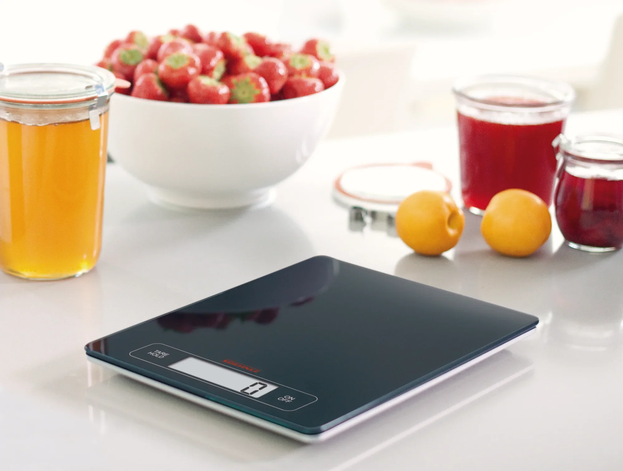 Page Profi digitale keukenweegschaal op een keukenblad, omringd door verse aardbeien in een schaal en potten jam en sap. Ideaal voor nauwkeurig wegen.