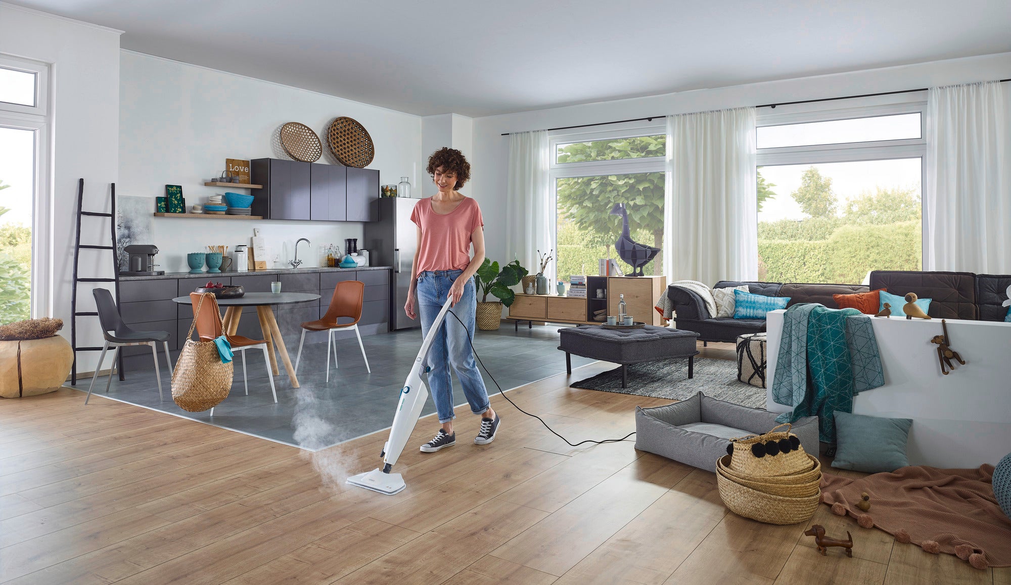 Eine Frau reinigt den Holzboden eines modernen Wohnzimmers mit dem Dampfreiniger CleanTenso Power. Der Raum ist hell und stilvoll eingerichtet, mit großen Fenstern und einer offenen Küche.