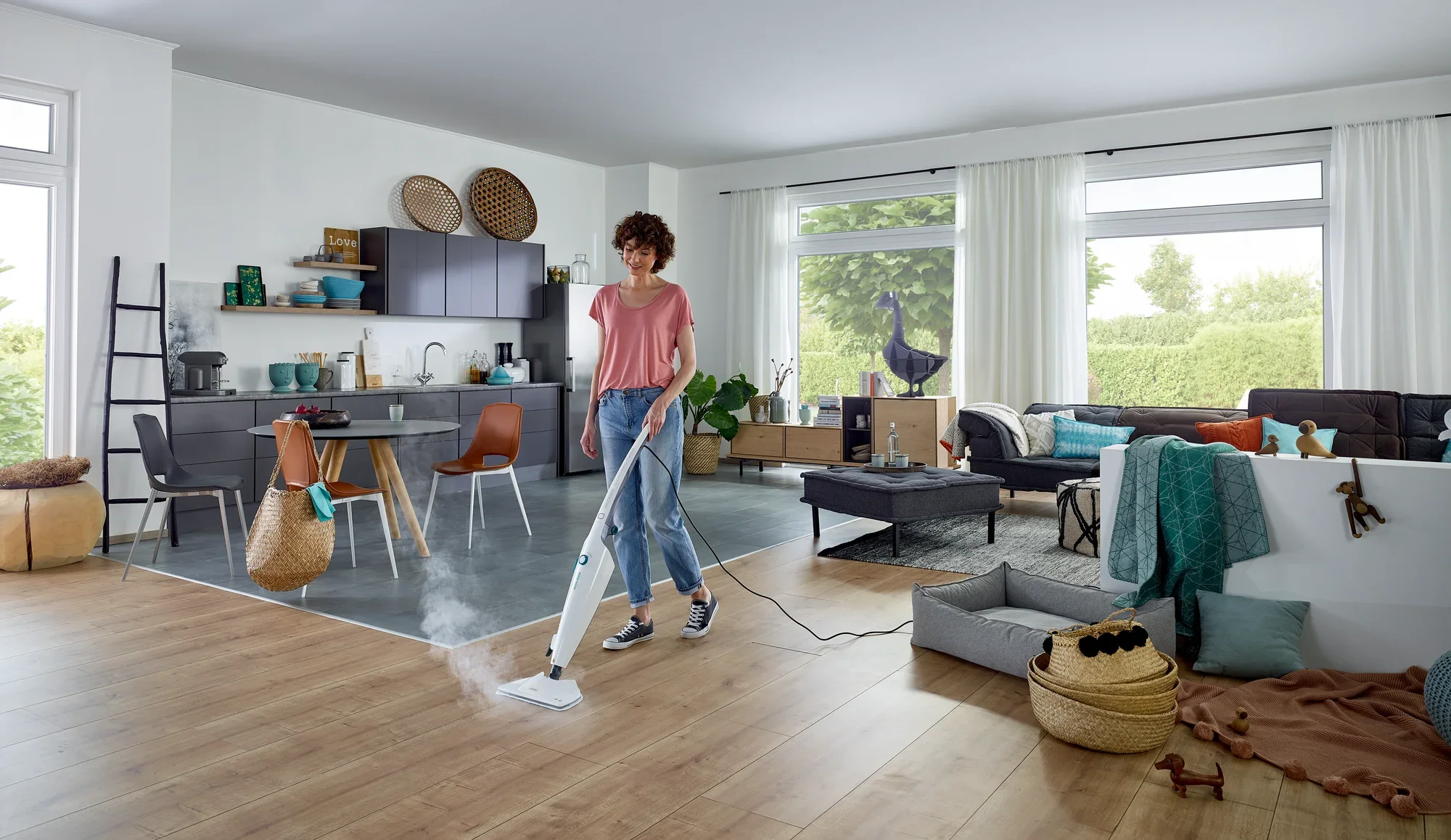 Une femme nettoie le sol en bois d'un salon moderne avec le nettoyeur vapeur CleanTenso Power. La pièce est lumineuse et élégamment meublée, avec de grandes fenêtres et une cuisine ouverte.