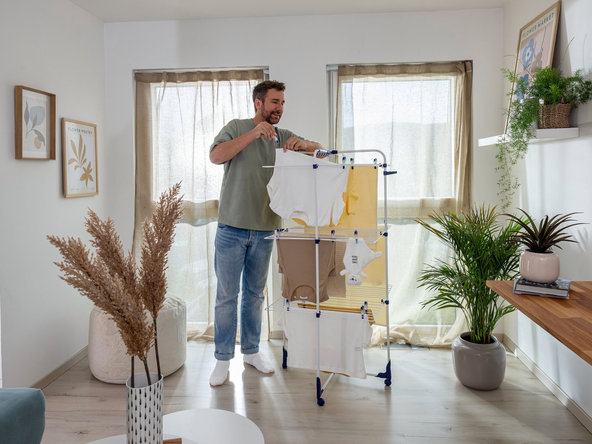 Een man staat in een lichte kamer en gebruikt de Classic Tower 340 wasdroger om de was te drogen. Aan de droger hangen verschillende kledingstukken. Planten en foto's sieren de kamer.