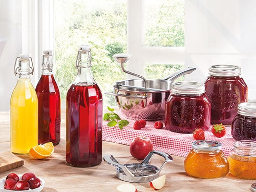 Verschiedene Flaschen und Glaeser mit roten und gelben Saeften und Marmeladen auf einem Tisch, umgeben von frischem Obst und einem Topf.