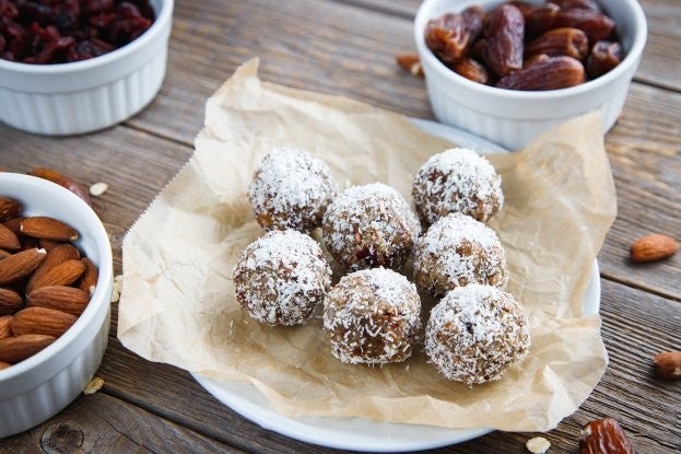 Hausgemachte Energyballs mit Kokosraspeln, Datteln und Mandeln auf Pergamentpapier, umgeben von Schalen mit Zutaten.