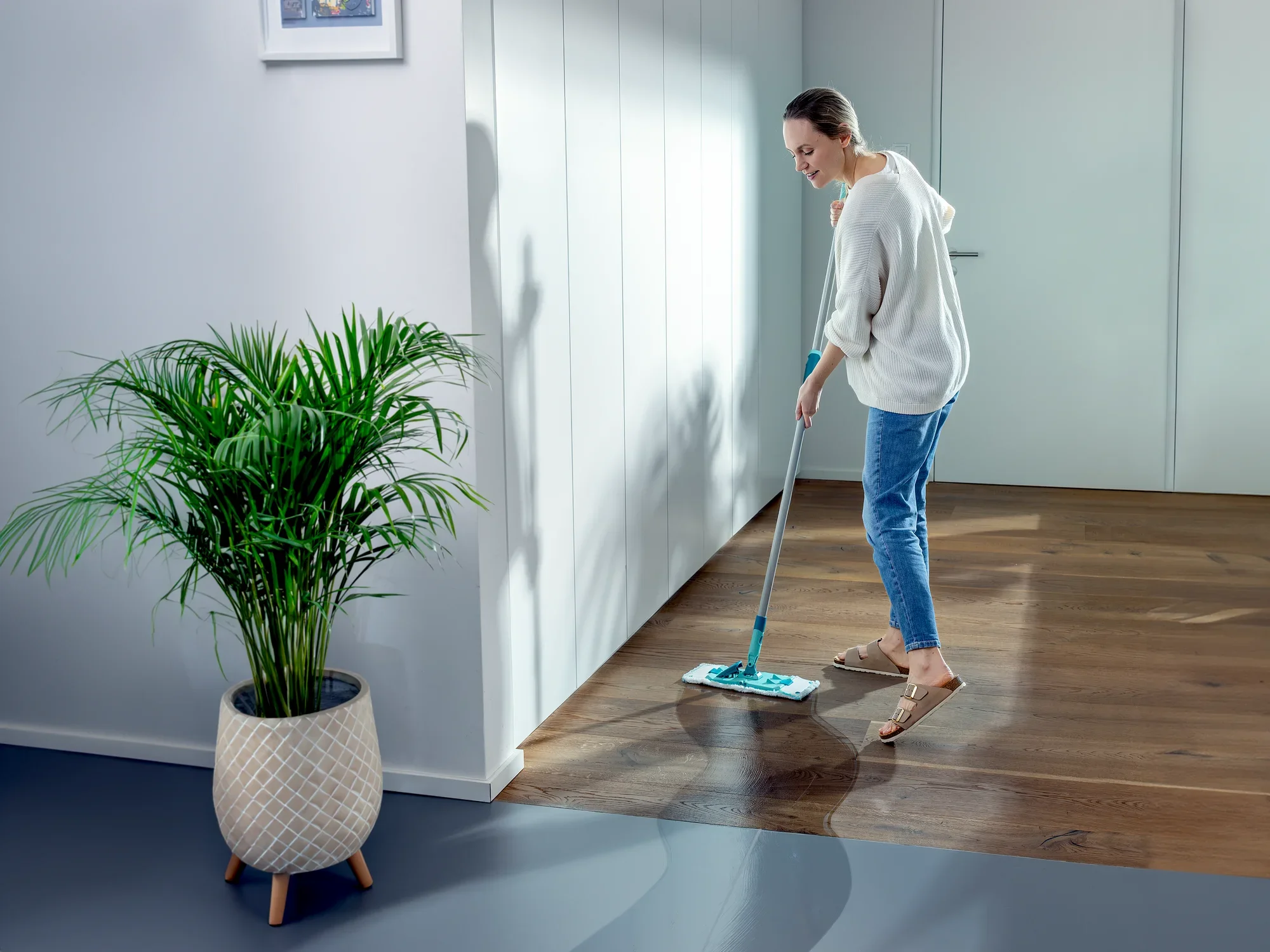 Une femme nettoie le sol en bois avec le balai POWER CLEAN M micro duo Telescope. Au premier plan, on voit une plante verte dans un pot décoratif. La scène montre un appartement lumineux et moderne.