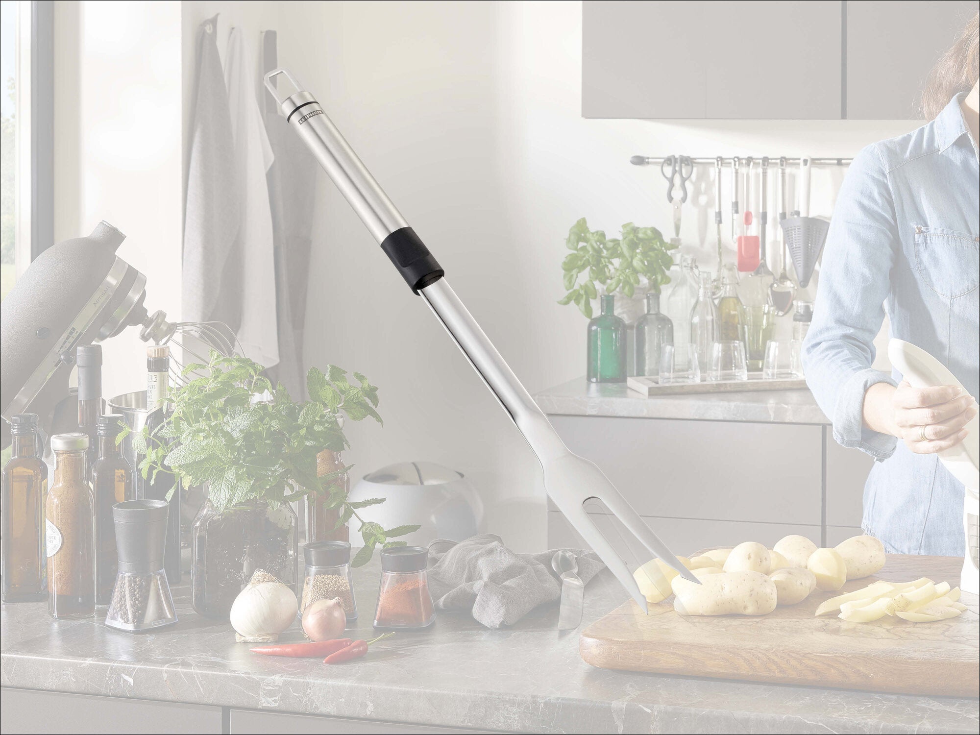 Leifheit Küchenrollenhalter Parat Royal aus silbernem Metall mit schwarzem Griff steht schräg auf der Arbeitsplatte in einer modernen Küche neben Kräutern, Flaschen und geschnittenen Kartoffeln.