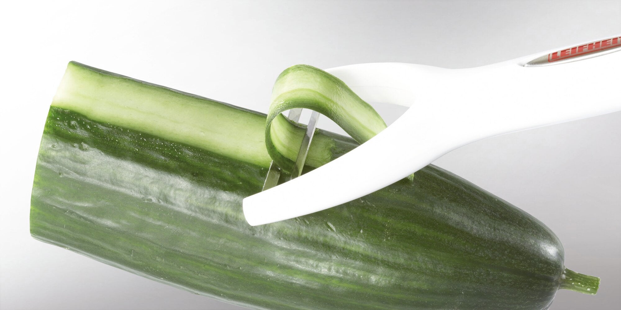 A white peeler efficiently removes a thin strip of green skin from a fresh cucumber, revealing a lighter interior.