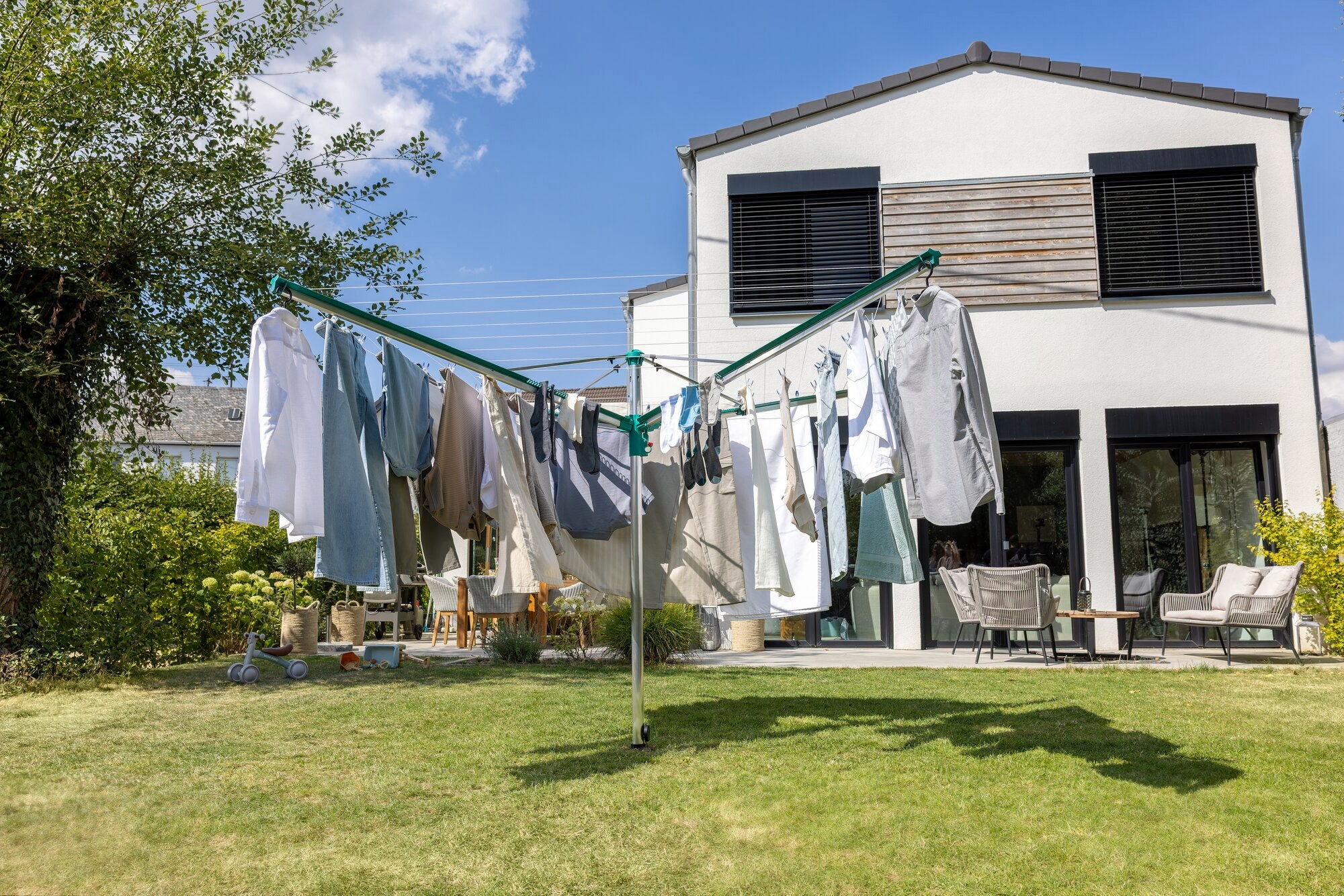 Wäschespinne Linomatic 500 Deluxe von Leifheit im Einsatz in einem sonnigen Garten. Die Wäschespinne ist mit verschiedenen Kleidungsstücken behängt, während im Hintergrund ein modernes Haus zu sehen ist.