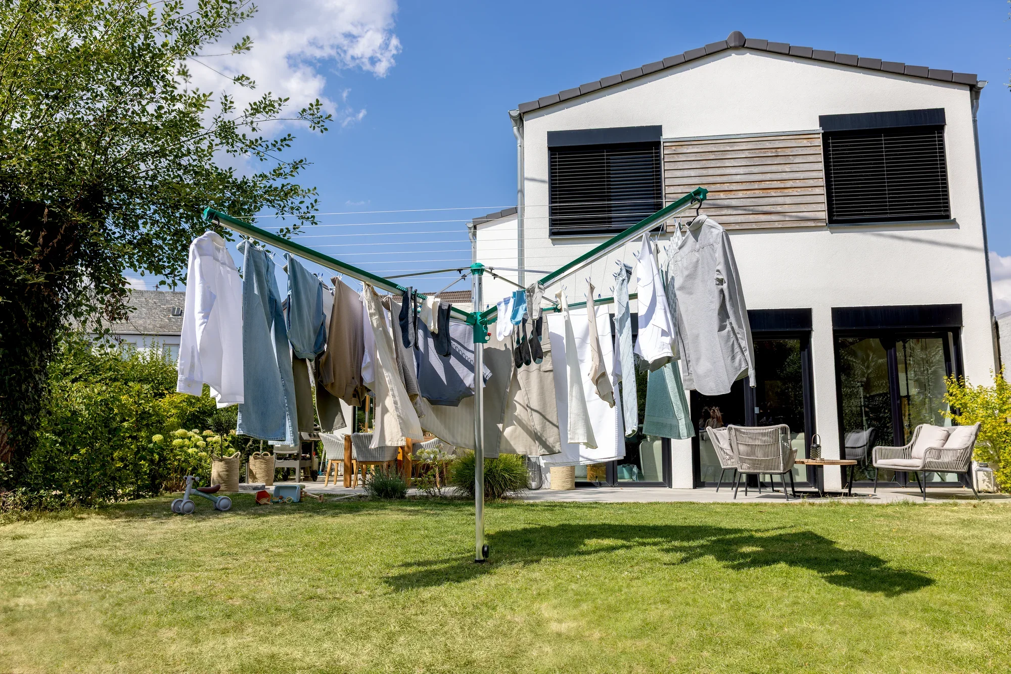 Wäschespinne Linomatic 500 Deluxe von Leifheit im Einsatz in einem sonnigen Garten. Die Wäschespinne ist mit verschiedenen Kleidungsstücken behängt, während im Hintergrund ein modernes Haus zu sehen ist.