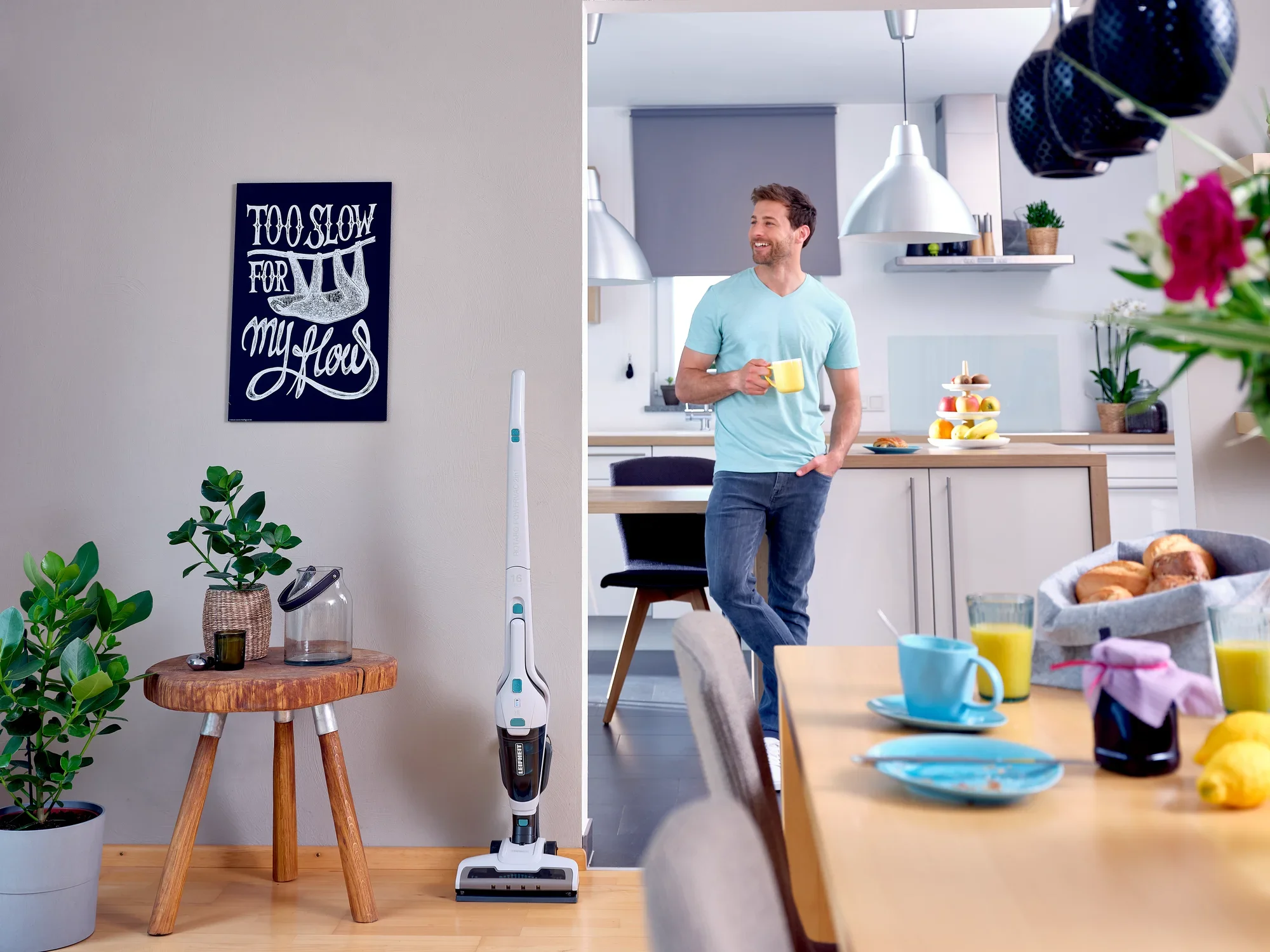 Un homme se tient dans une cuisine moderne avec un aspirateur sans fil Rotaro PowerVac. La pièce est lumineuse et accueillante, avec des plantes et une table dressée au premier plan.