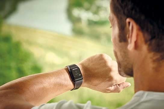 Ein Mann blickt auf eine Smartwatch an seinem Handgelenk, waehrend er in eine gruene Landschaft schaut.