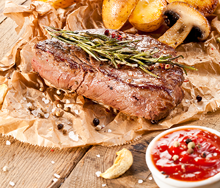Gegrilltes Rumpsteak mit Rosmarin auf Papier, daneben geroestete Kartoffeln, Champignons und ein Schaelchen mit roter Sosse auf Holztisch.