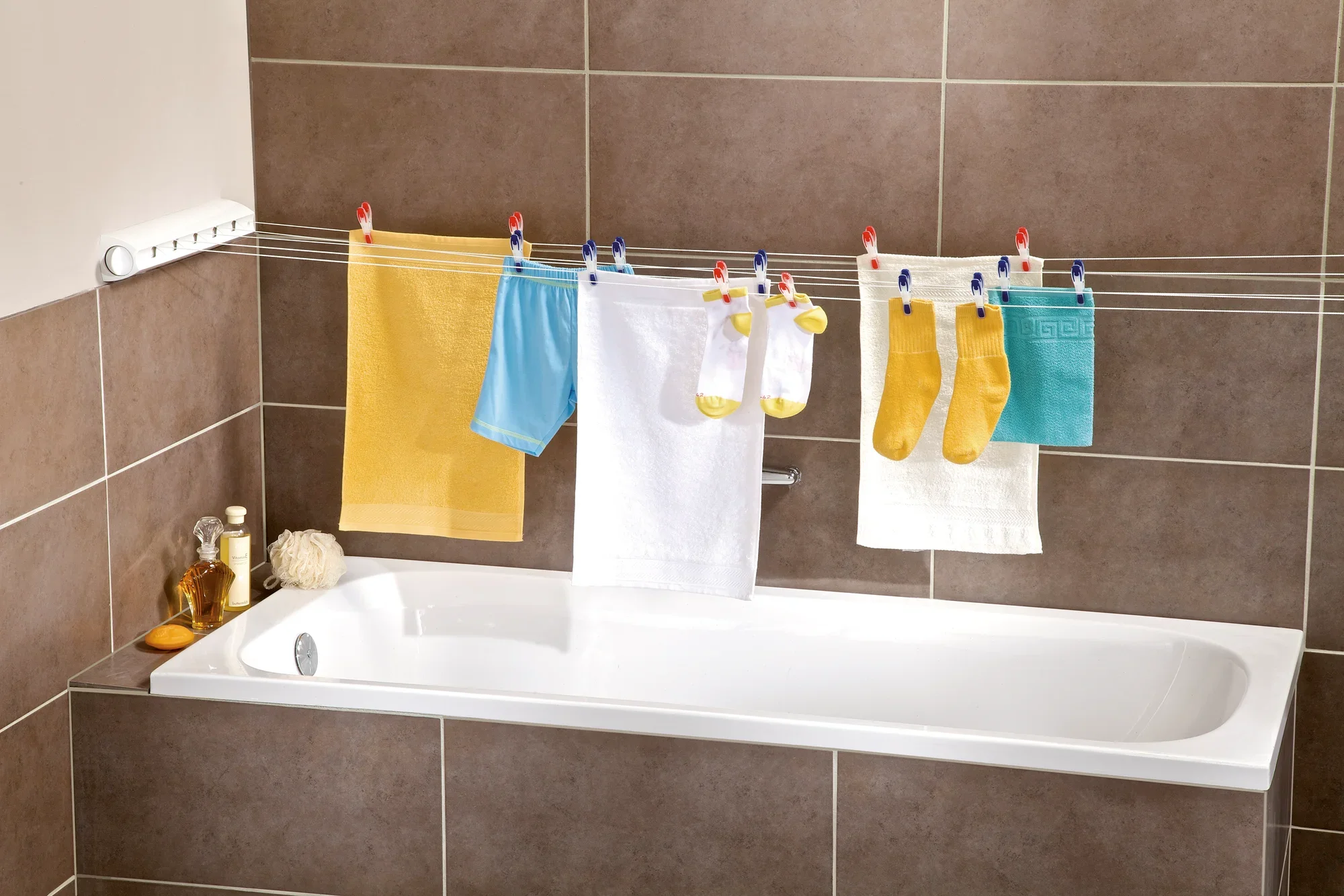 Suszarka ścienna Rollfix 210 Longline zamontowana nad wanną, z kilkoma kolorowymi ręcznikami i skarpetkami wiszącymi do wyschnięcia. Idealna do suszenia oszczędzającego miejsce.