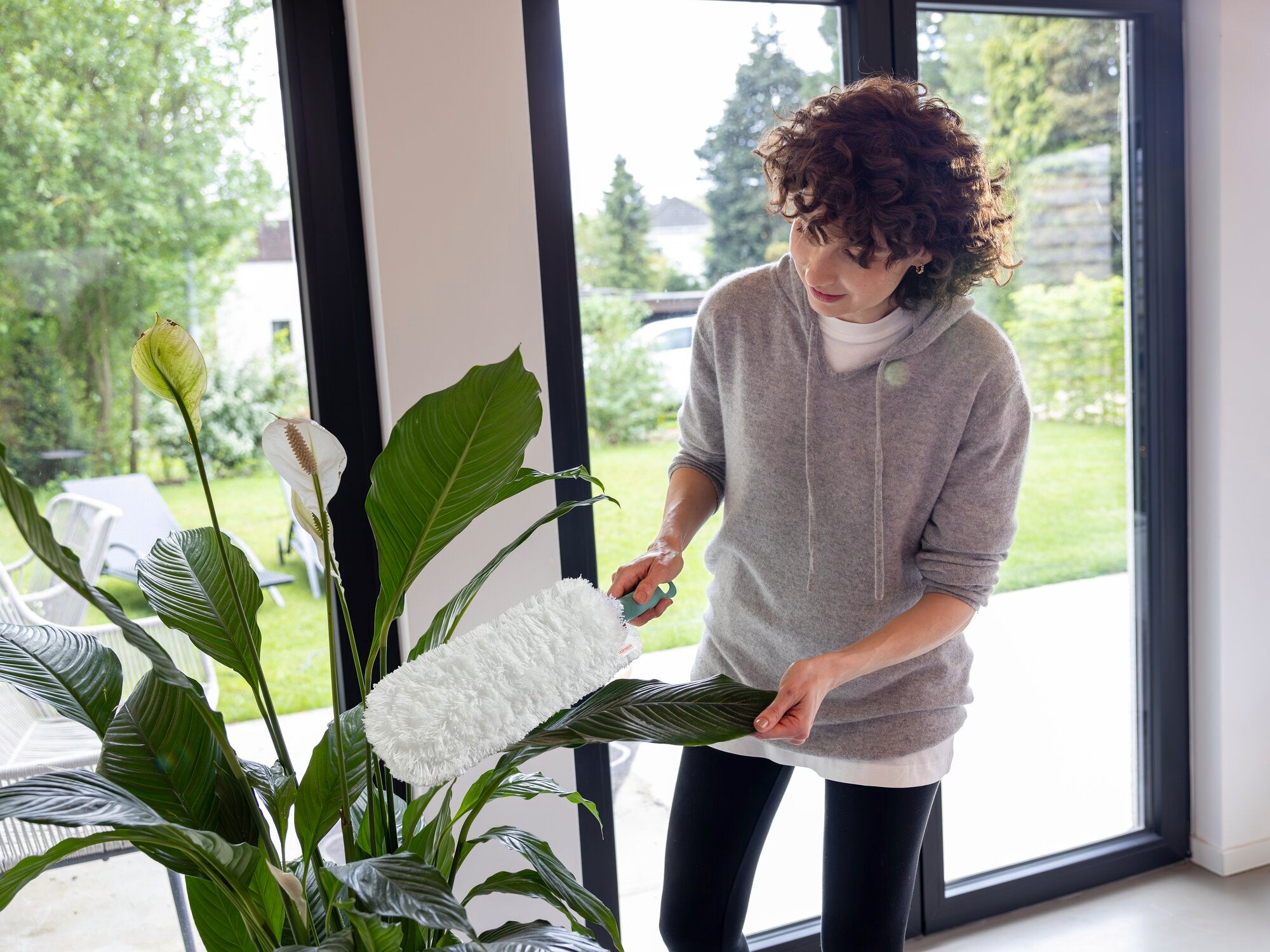 Een persoon gebruikt de SUPERDUSTER XL plumeau om stof te verwijderen van de grote groene bladeren van een kamerplant. Op de achtergrond is een lichte kamer met grote ramen te zien.