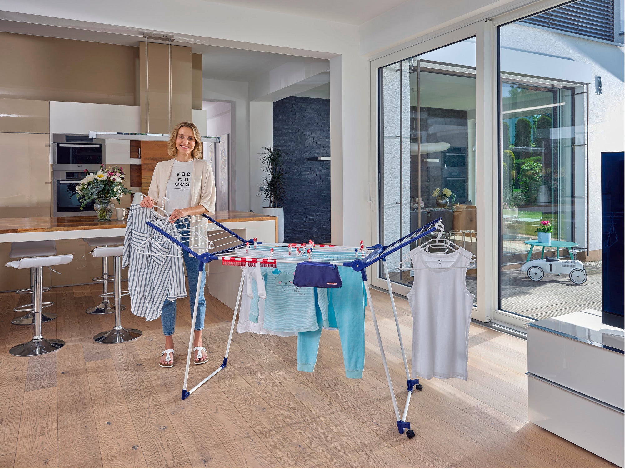 Eine Frau steht in einem modernen Wohnzimmer und benutzt den Standtrockner Pegasus 200 Solid Deluxe Mobile. Der Trockner ist mit Kleidung beladen und steht vor großen Fenstern.