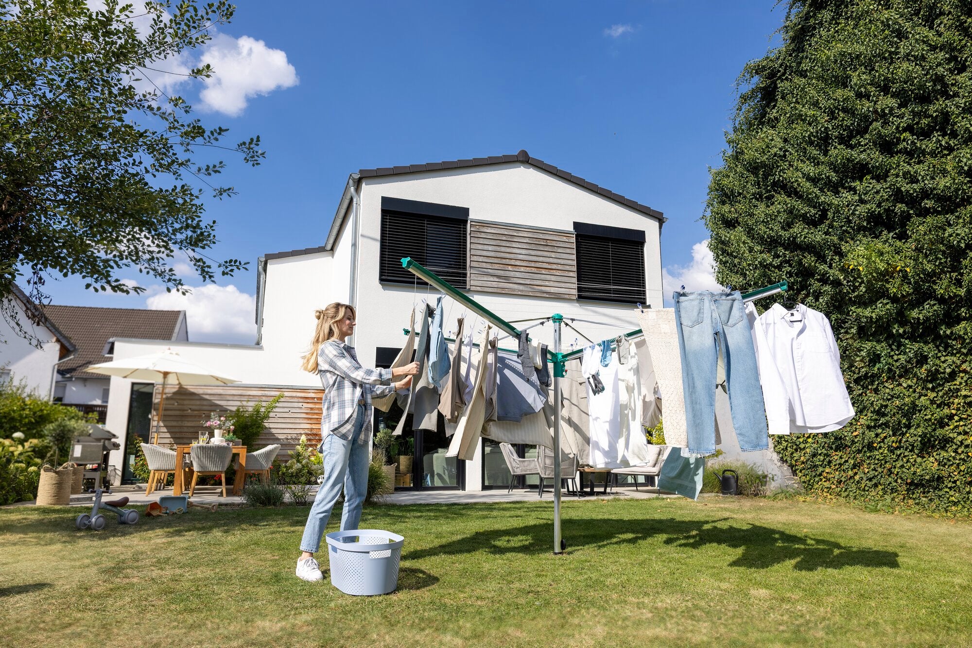 Eine Frau hängt Wäsche auf die Leifheit Wäschespinne Linomatic 500 Deluxe in einem sonnigen Garten. Im Hintergrund steht ein modernes Haus mit Terrasse und Gartenmöbeln.