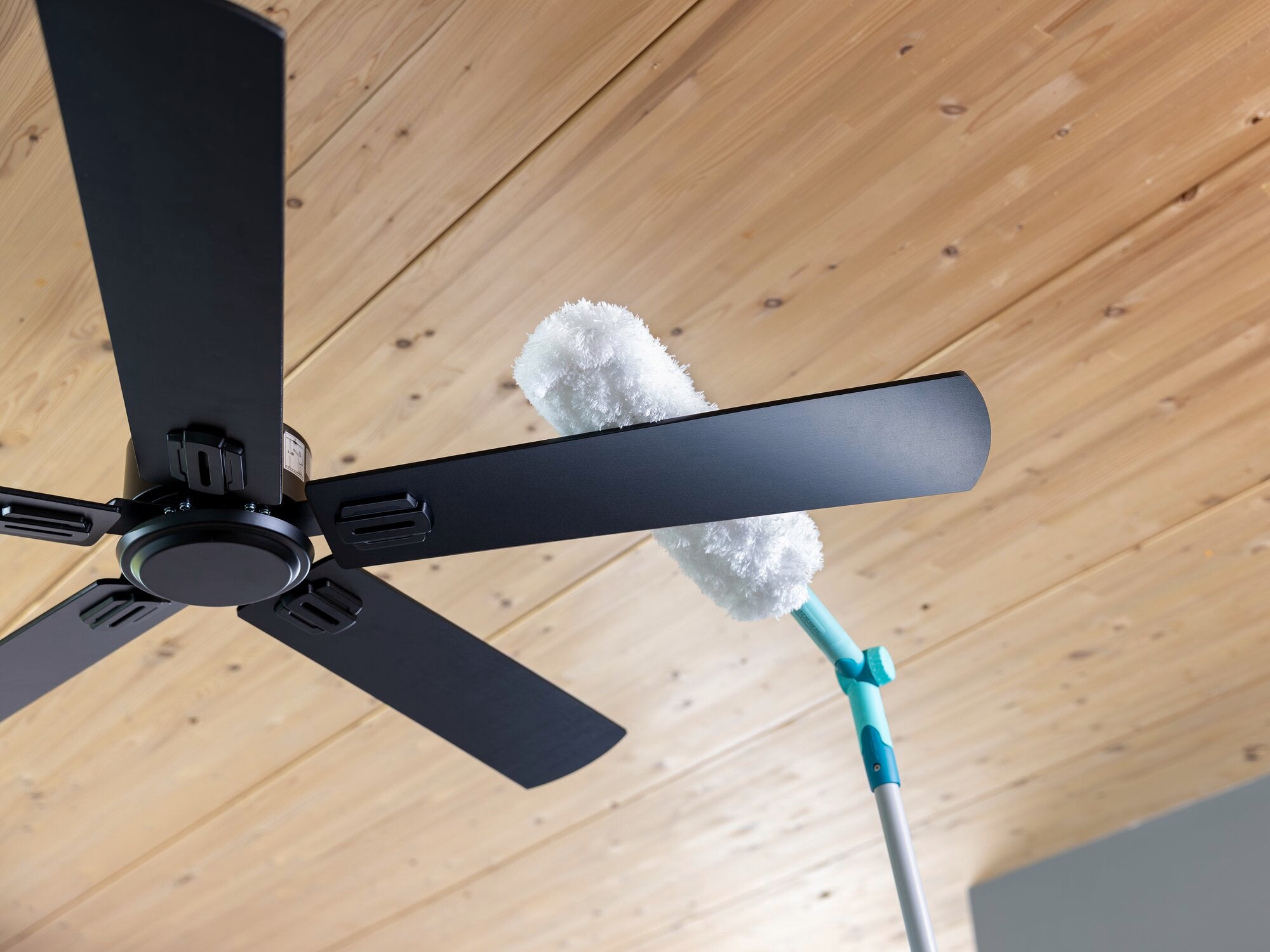 Ein SUPERDUSTER XL Pro Reach Staubwedel reinigt die Flügel eines Deckenventilators. Der Staubwedel hat einen langen Griff, ideal für schwer erreichbare Stellen.