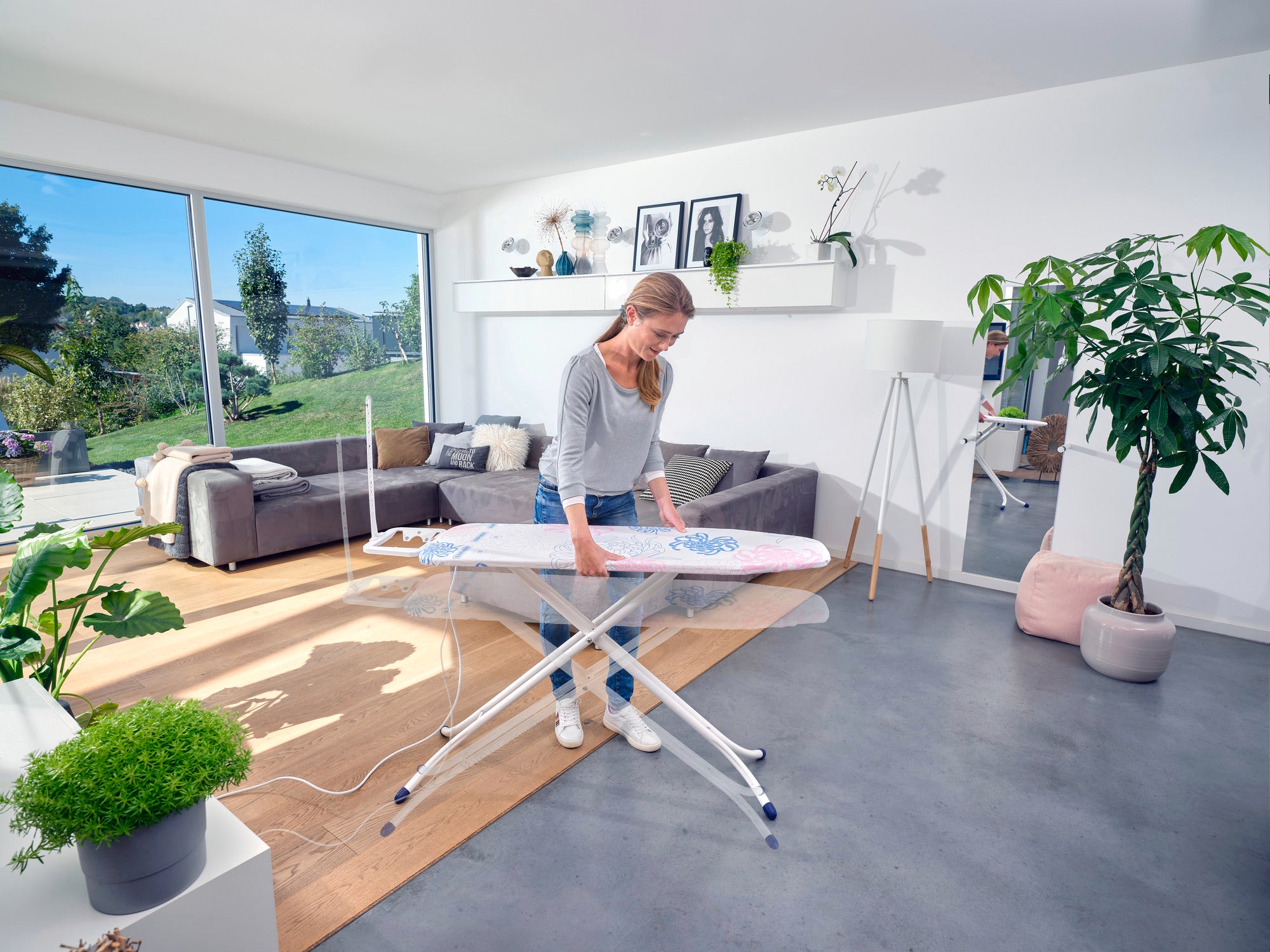 Bügeltisch Classic M Compact Plus in einem modernen Wohnzimmer. Eine Person bügelt auf dem Tisch, umgeben von Pflanzen und stilvoller Einrichtung. Große Fenster bieten Blick ins Freie.