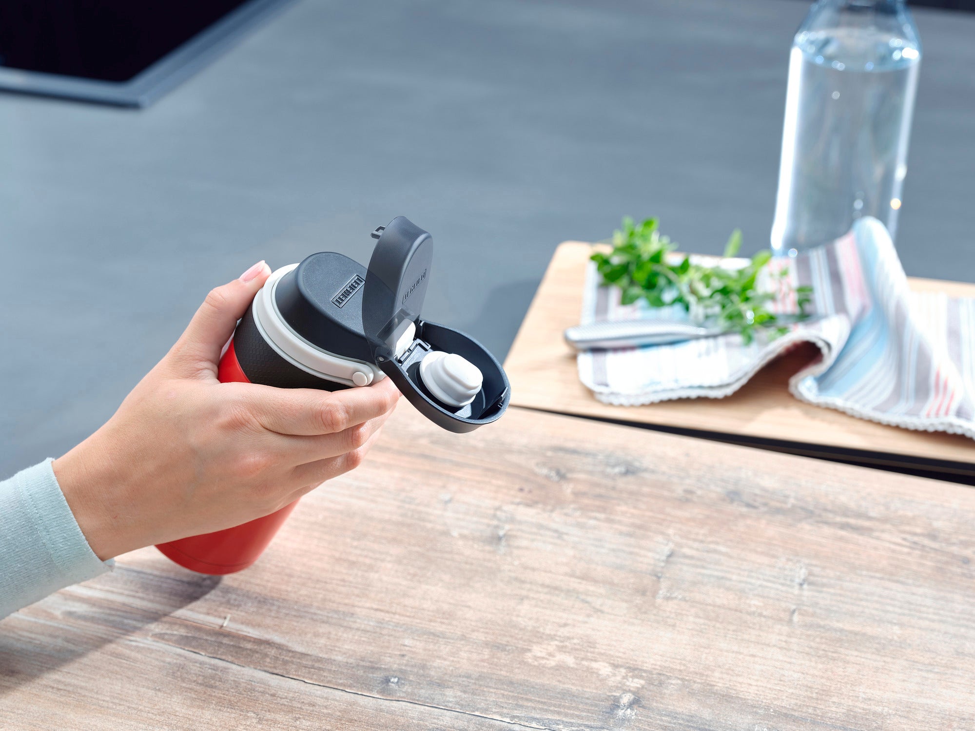 Une personne tient un mug isotherme Flip rouge d'une contenance de 350 ml. Le mug est équipé d'un couvercle rabattable pratique. À l'arrière-plan, on aperçoit une table, une serviette et une bouteille d'eau.