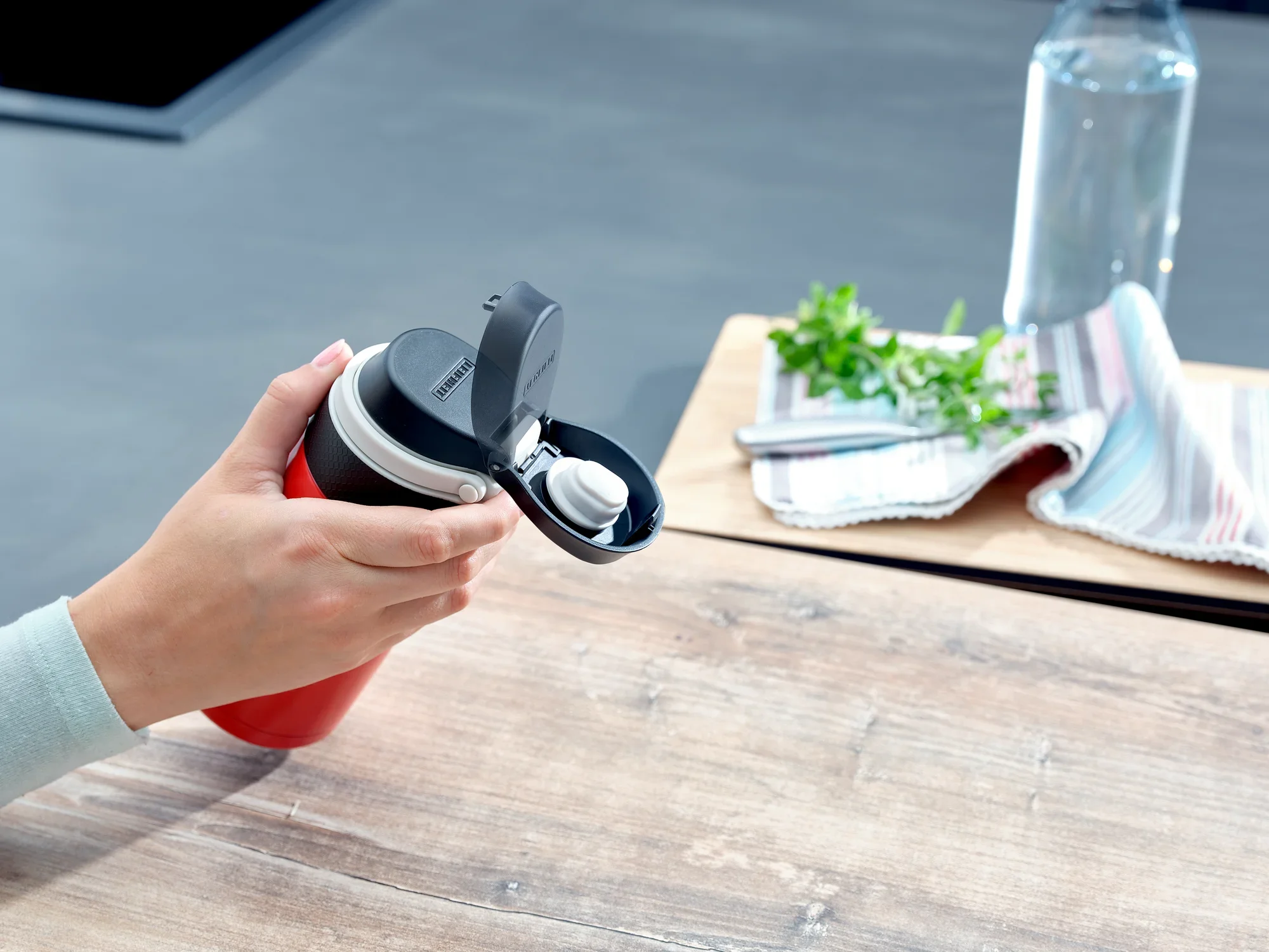 Jedna osoba trzyma czerwony izolowany kubek Flip o pojemności 350 ml. Kubek ma praktyczną pokrywkę typu flip-top. W tle widać stół, ręcznik i butelkę z wodą.