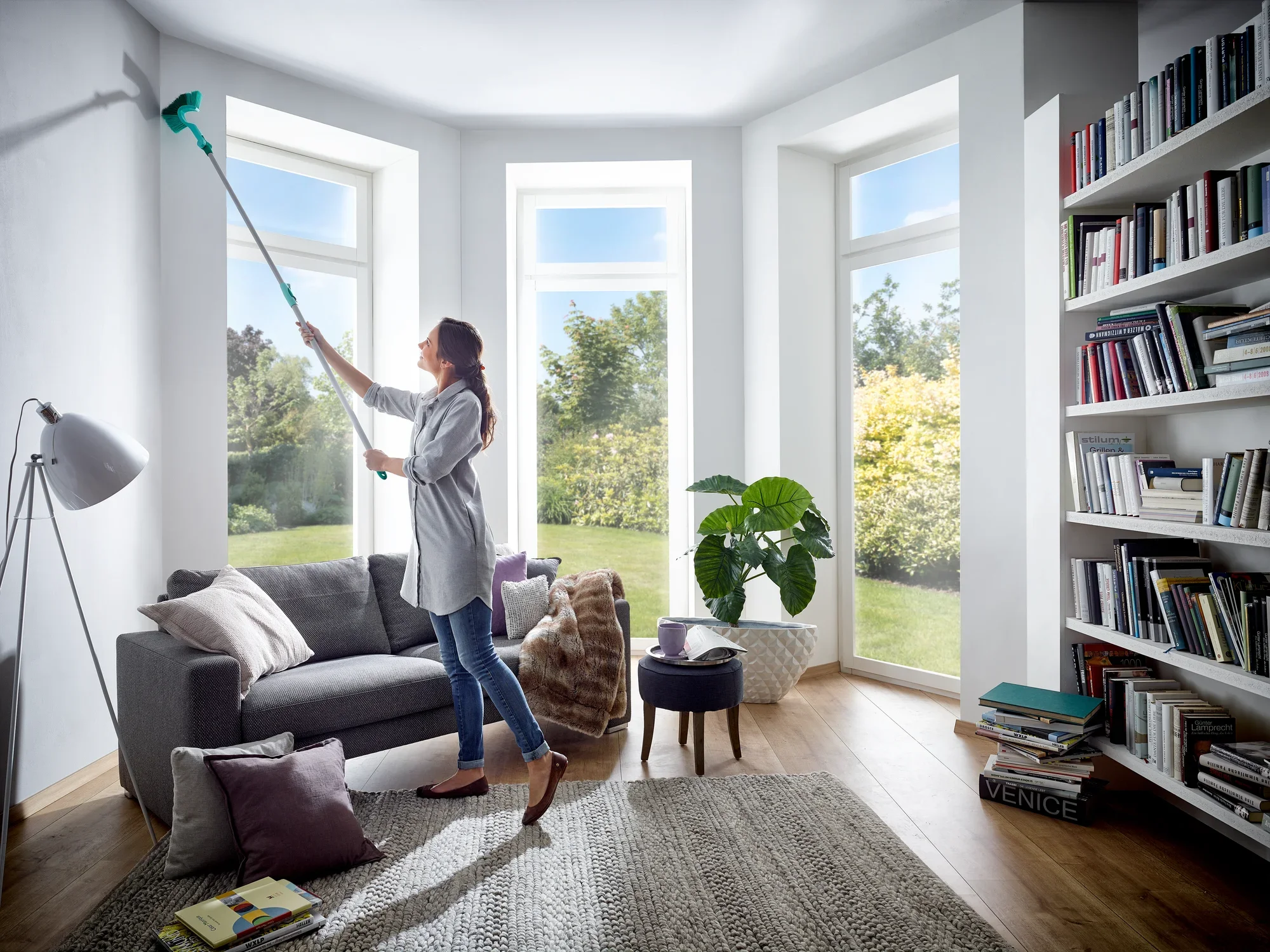 Een vrouw gebruikt de stofbezem met telescopische steel om hoge hoeken van een lichte woonkamer schoon te maken. Grote ramen laten veel licht binnen en rechts staat een boekenplank.