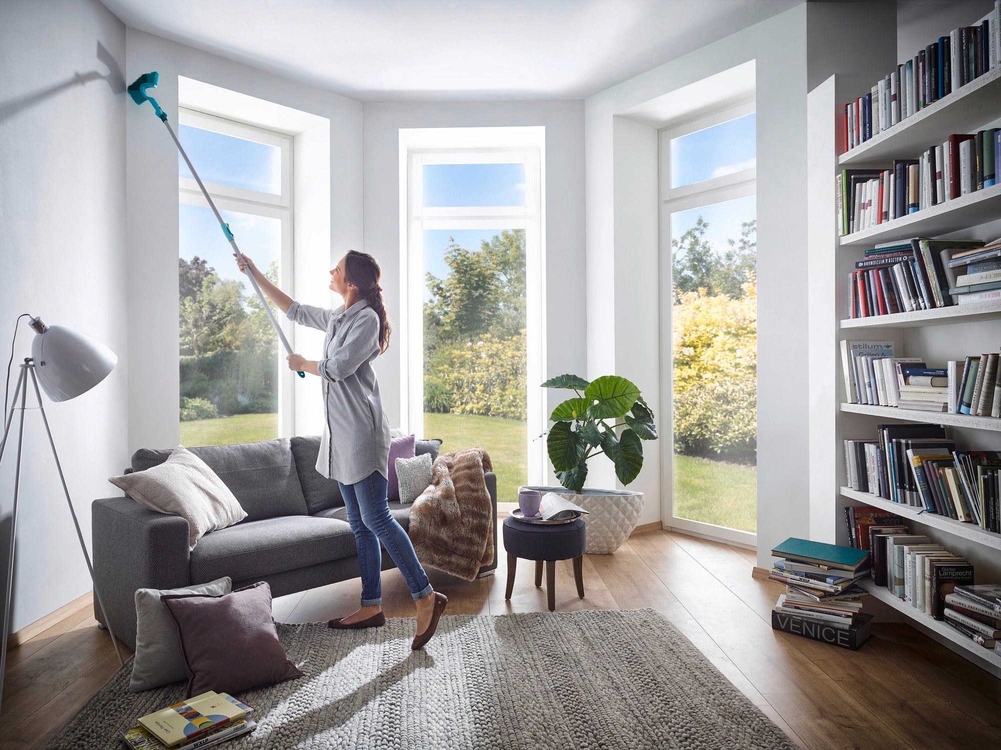 A woman uses the dusting brush with telescopic handle to clean high corners in a bright living room. Large windows let in plenty of light, and there is a bookcase on the right.