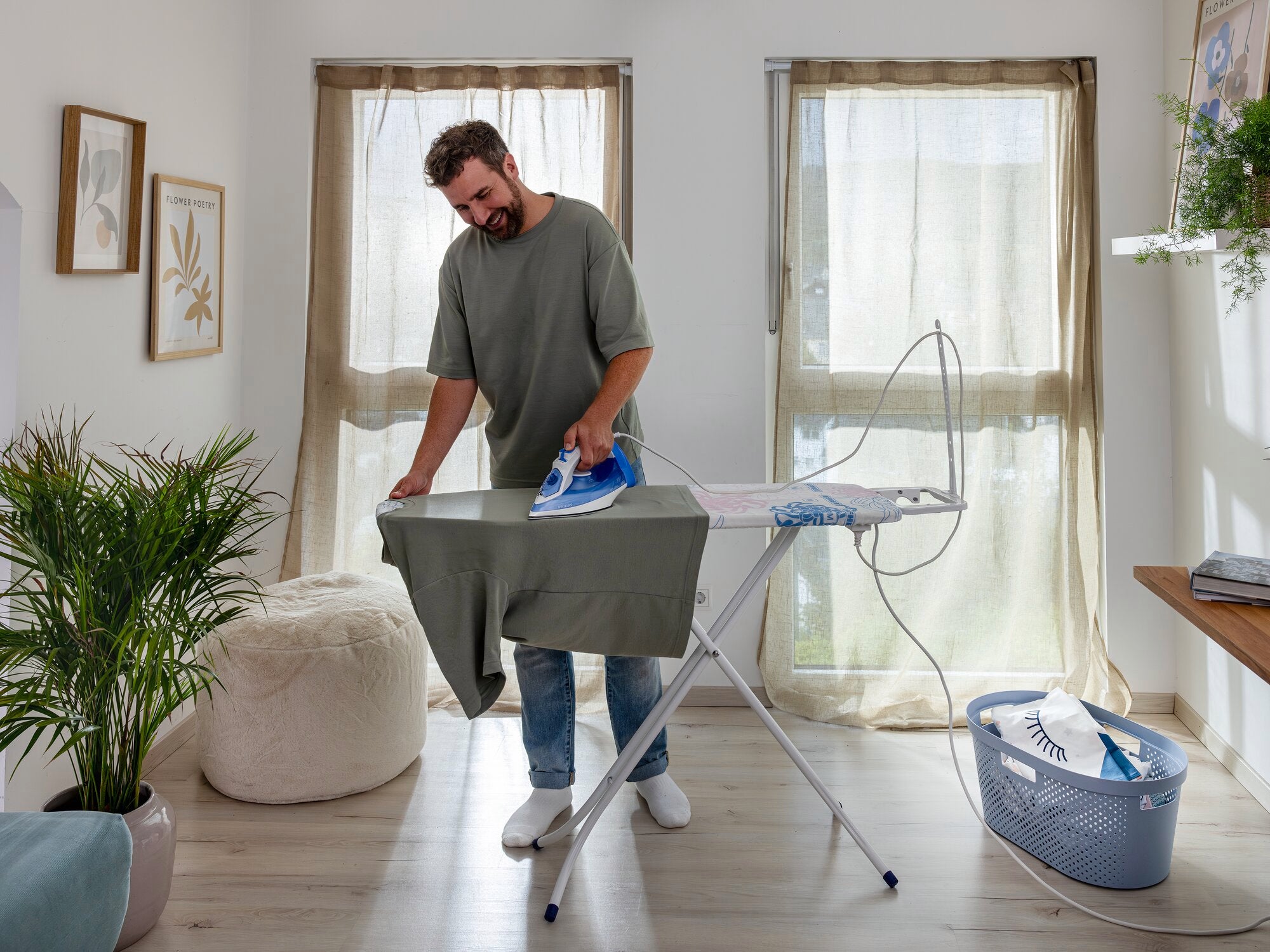 Un homme repasse une chemise sur la table à repasser Classic M Basic Plus dans une pièce lumineuse. La table se trouve devant deux fenêtres, à côté d'un panier à linge.