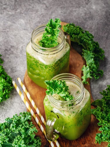 Zwei Glaeser mit gruenem Gruenkohl-Smoothie, garniert mit Gruenkohlblaettern, stehen auf einem Holzbrett, umgeben von frischen Gruenkohlblaettern.