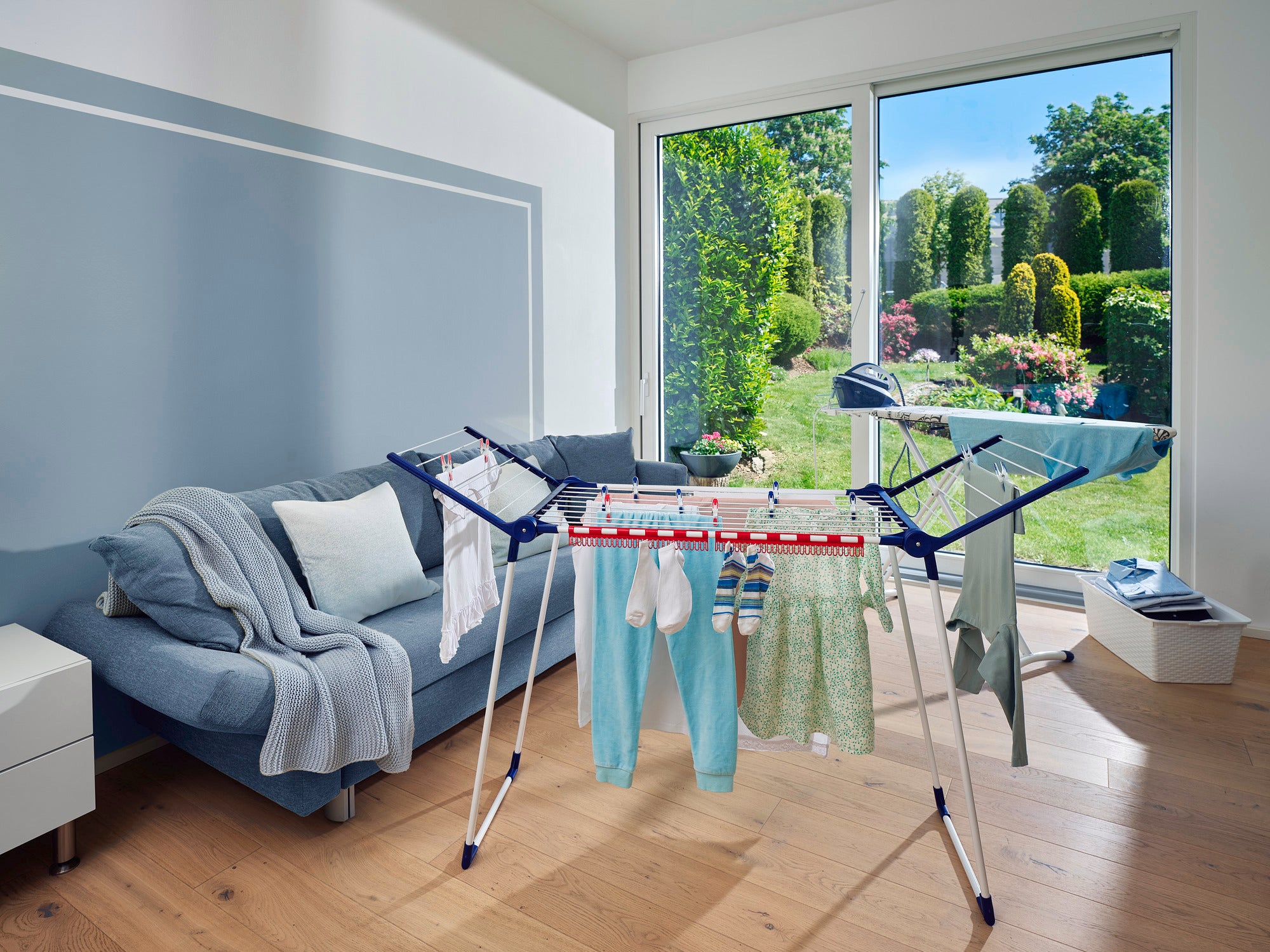Ein Standtrockner Pegasus 150 Solid Slim steht in einem modernen Wohnzimmer mit Blick auf einen sonnigen Garten. Der Trockner ist mit Kleidung beladen und bietet eine platzsparende Lösung.