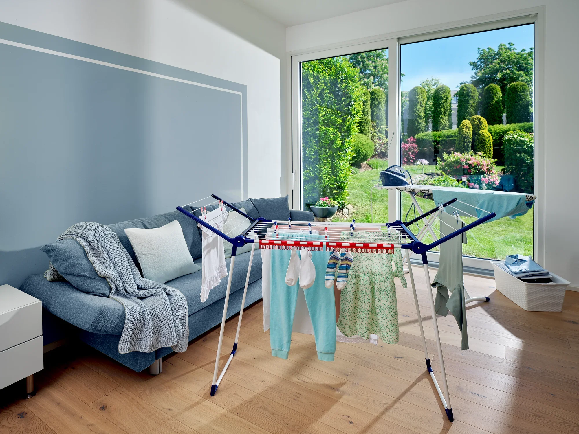 Ein Standtrockner Pegasus 150 Solid Slim steht in einem modernen Wohnzimmer mit Blick auf einen sonnigen Garten. Der Trockner ist mit Kleidung beladen und bietet eine platzsparende Lösung.