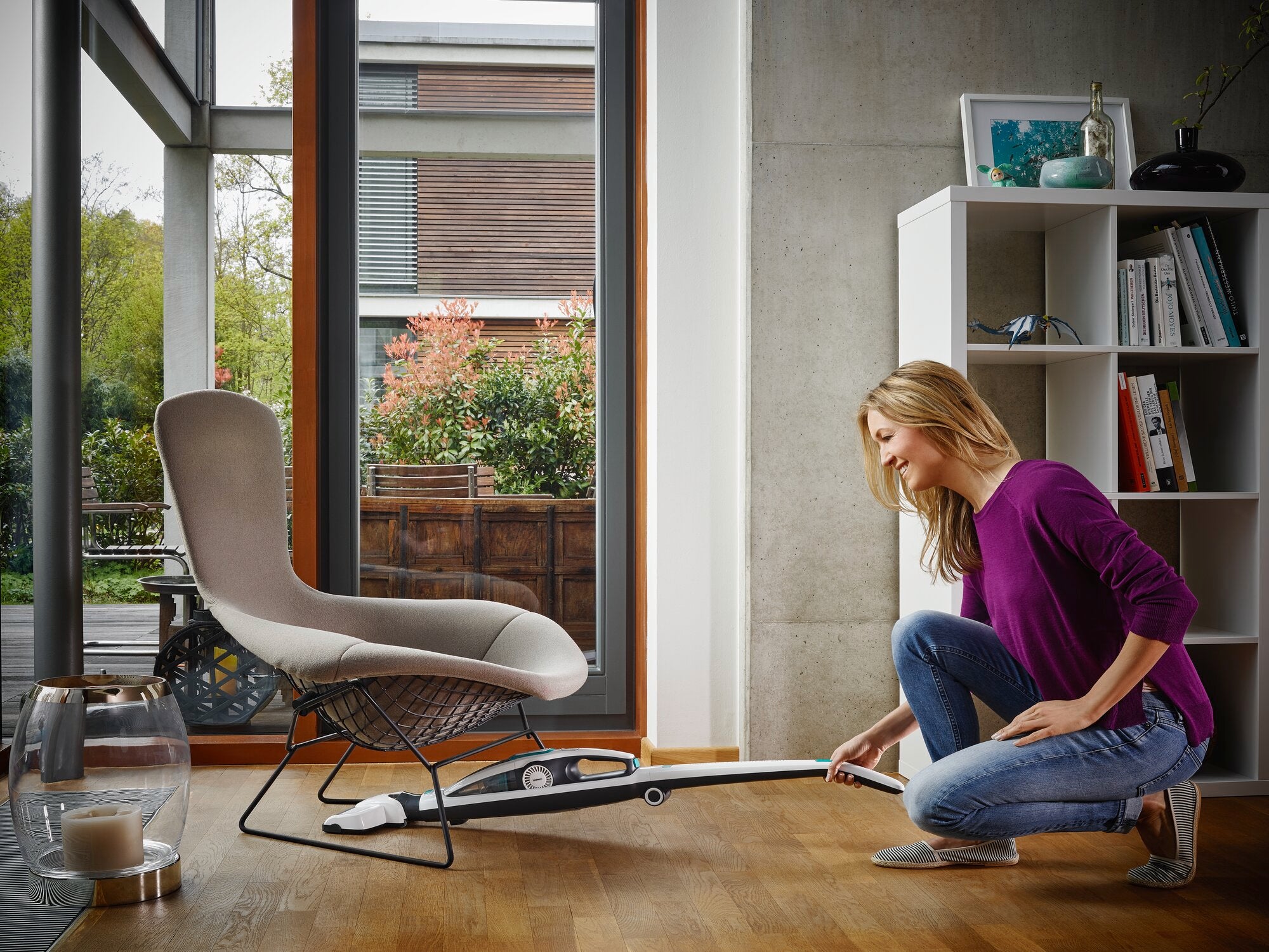 Eine Frau benutzt den Regulus PowerVac 2in1 Staubsauger, um den Boden unter einem modernen Stuhl in einem hellen Wohnzimmer zu reinigen. Der Staubsauger ist leicht und handlich.