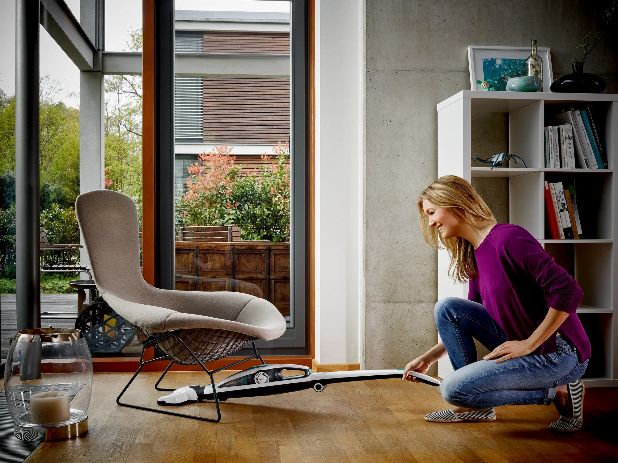Une femme utilise l'aspirateur Regulus PowerVac 2 en 1 pour nettoyer le sol sous une chaise moderne dans un salon lumineux. L'aspirateur est léger et maniable.