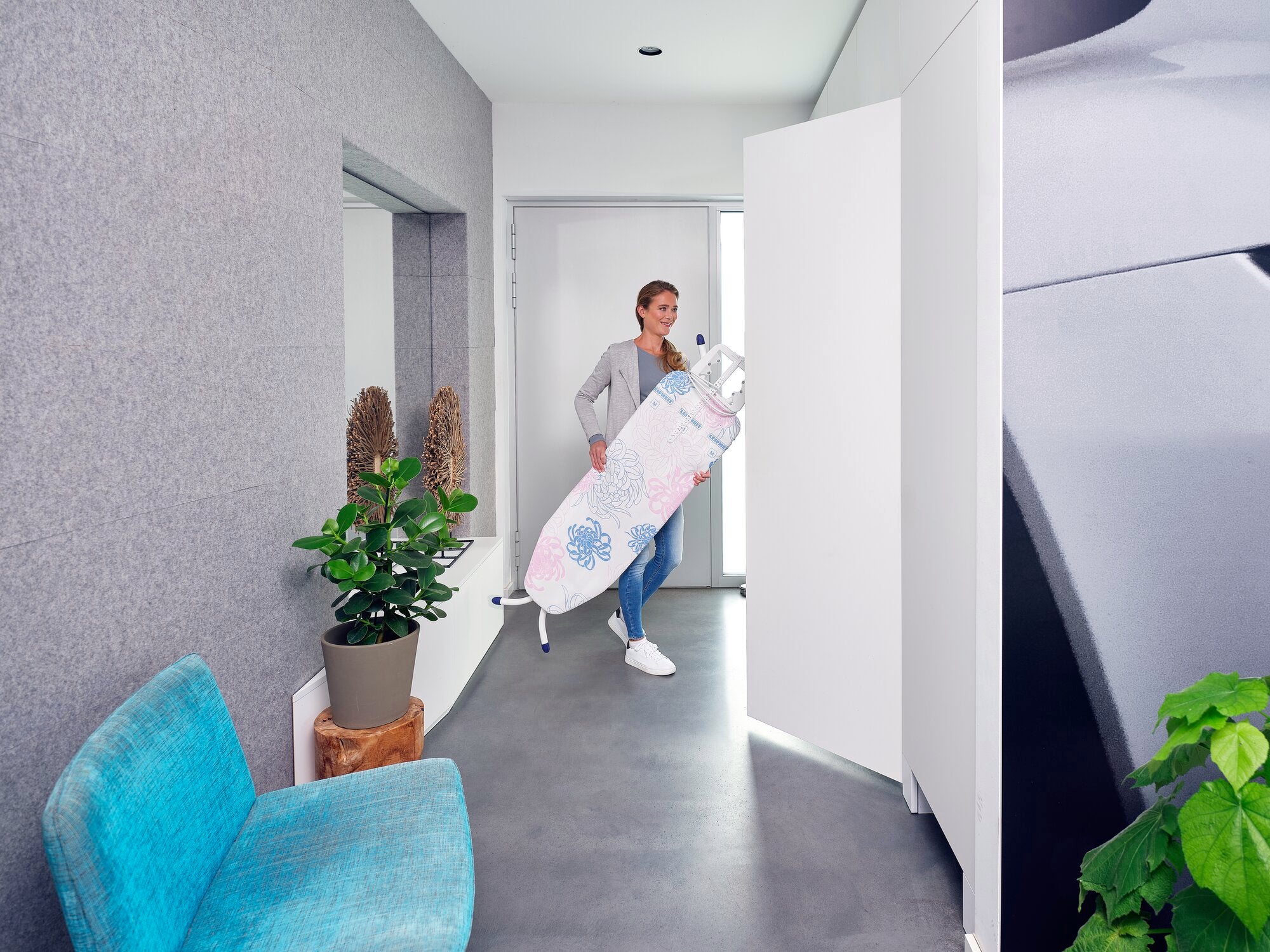 Bügelbrett Classic M Compact Plus: Eine Person trägt ein zusammengeklapptes Bügelbrett durch einen modernen, hellen Raum. Das Brett hat ein florales Muster und ist leicht zu transportieren.