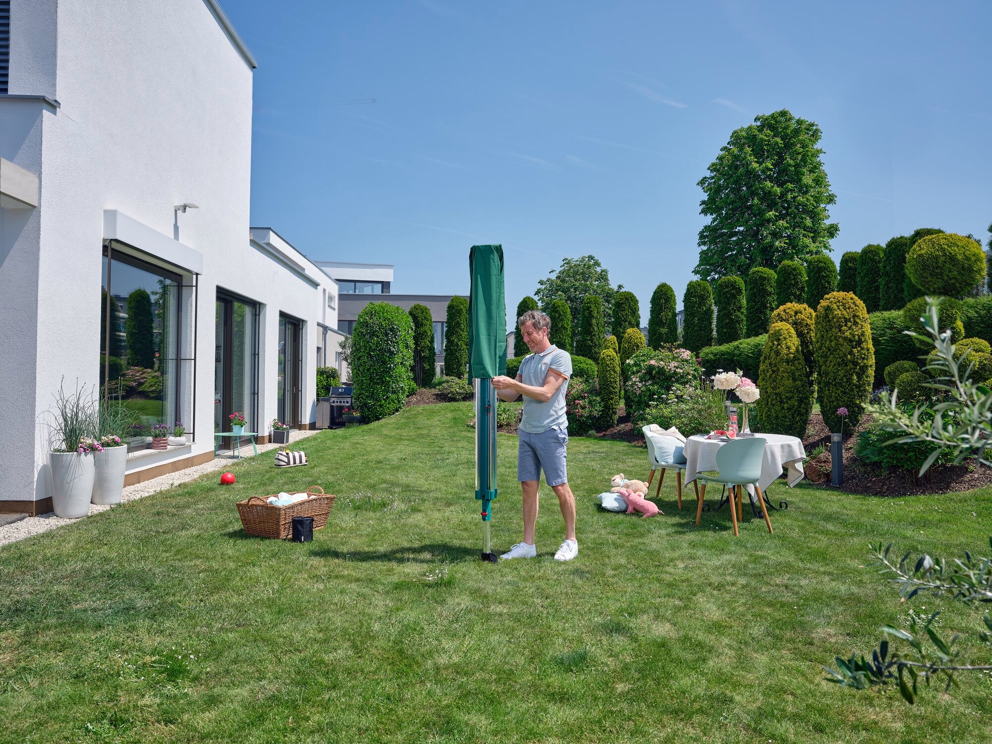 Ein Mann steht in einem gepflegten Garten neben einem modernen Haus und benutzt den Wäscheschirm Linomatic 600 Deluxe. Der Schirm ist grün und umgeben von Büschen und einem Tisch mit Blumen.