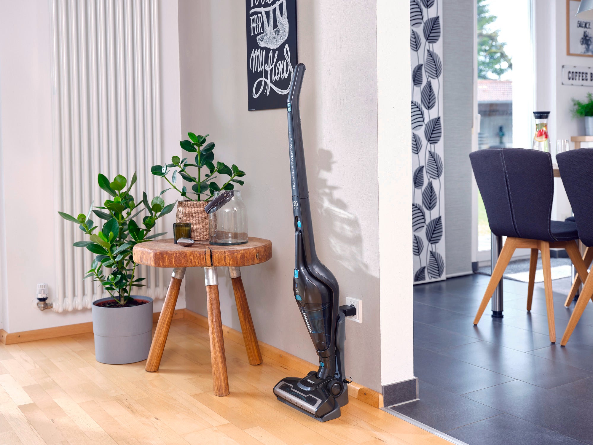 Ein Akku-Staubsauger Rotaro PowerVac steht in einem modernen Wohnzimmer neben einem Holztisch mit Pflanzen. Der Raum ist hell und stilvoll eingerichtet.
