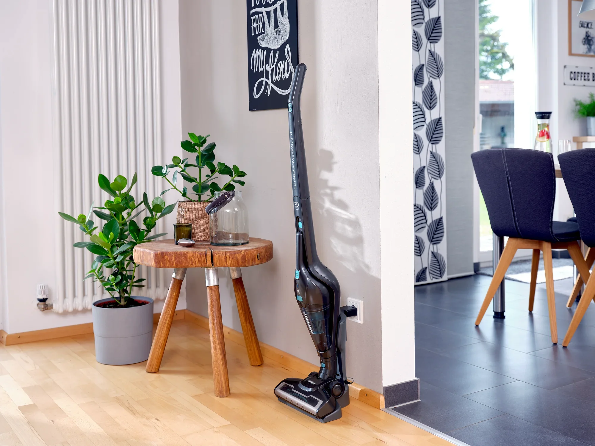 Un aspirateur sans fil Rotaro PowerVac se trouve dans un salon moderne à côté d'une table en bois avec des plantes. La pièce est lumineuse et meublée avec style.