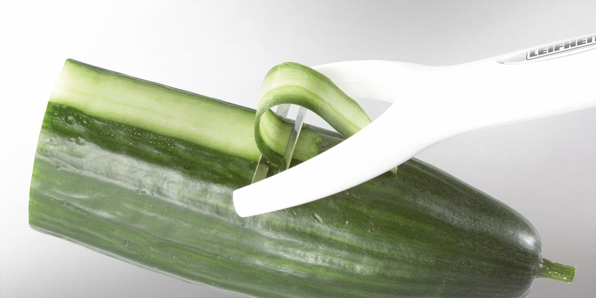 Ein weißer Leifheit Sparschäler schält eine Gurke. Der Schäler entfernt eine dünne Schicht der Schale, während die Gurke auf einer glatten Oberfläche liegt.