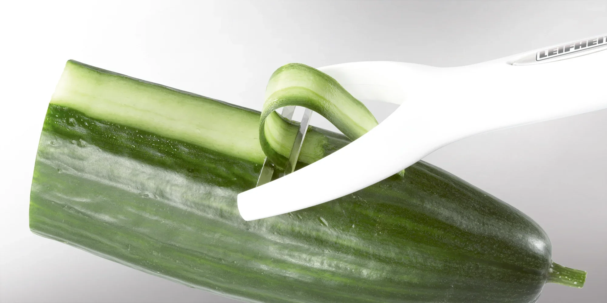 Ein weißer Leifheit Sparschäler schält eine Gurke. Der Schäler entfernt eine dünne Schicht der Schale, während die Gurke auf einer glatten Oberfläche liegt.