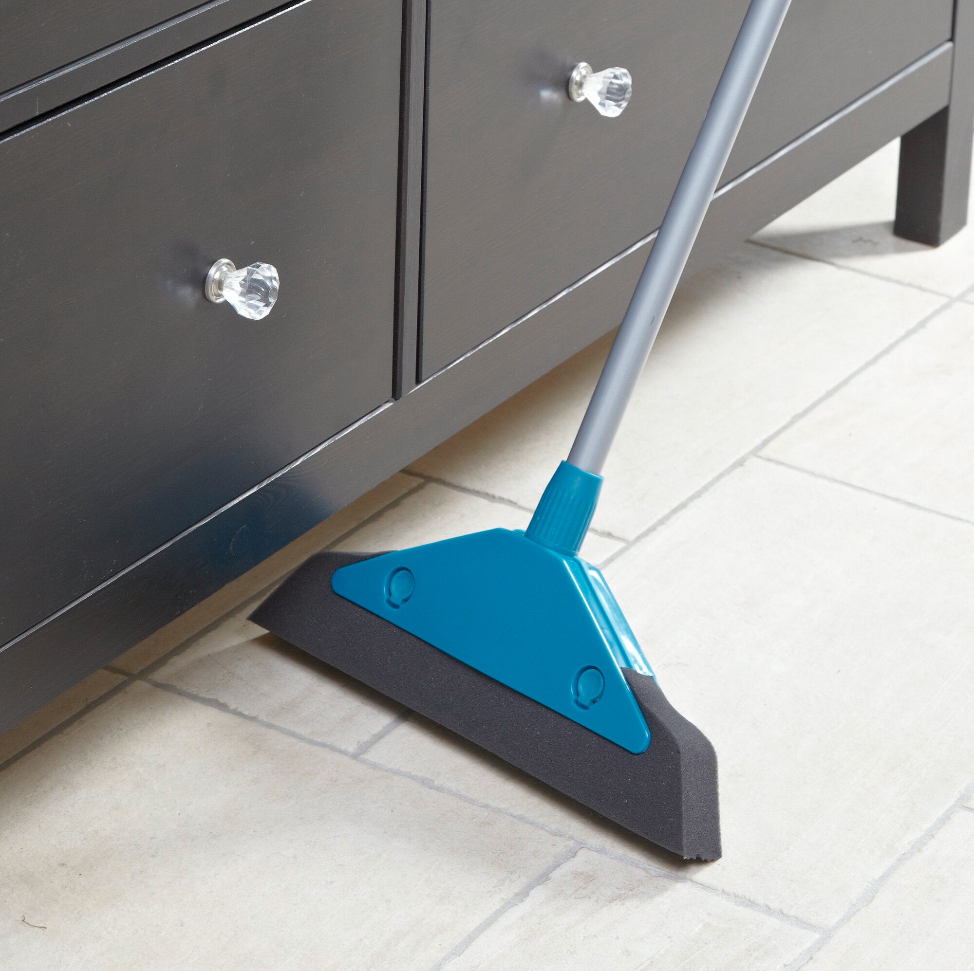 Ein blauer Schaumstoffbesen reinigt effektiv den Boden unter einem schwarzen Schrank mit glänzenden Griffen. Der Besen ist ideal für schwer zugängliche Stellen.
