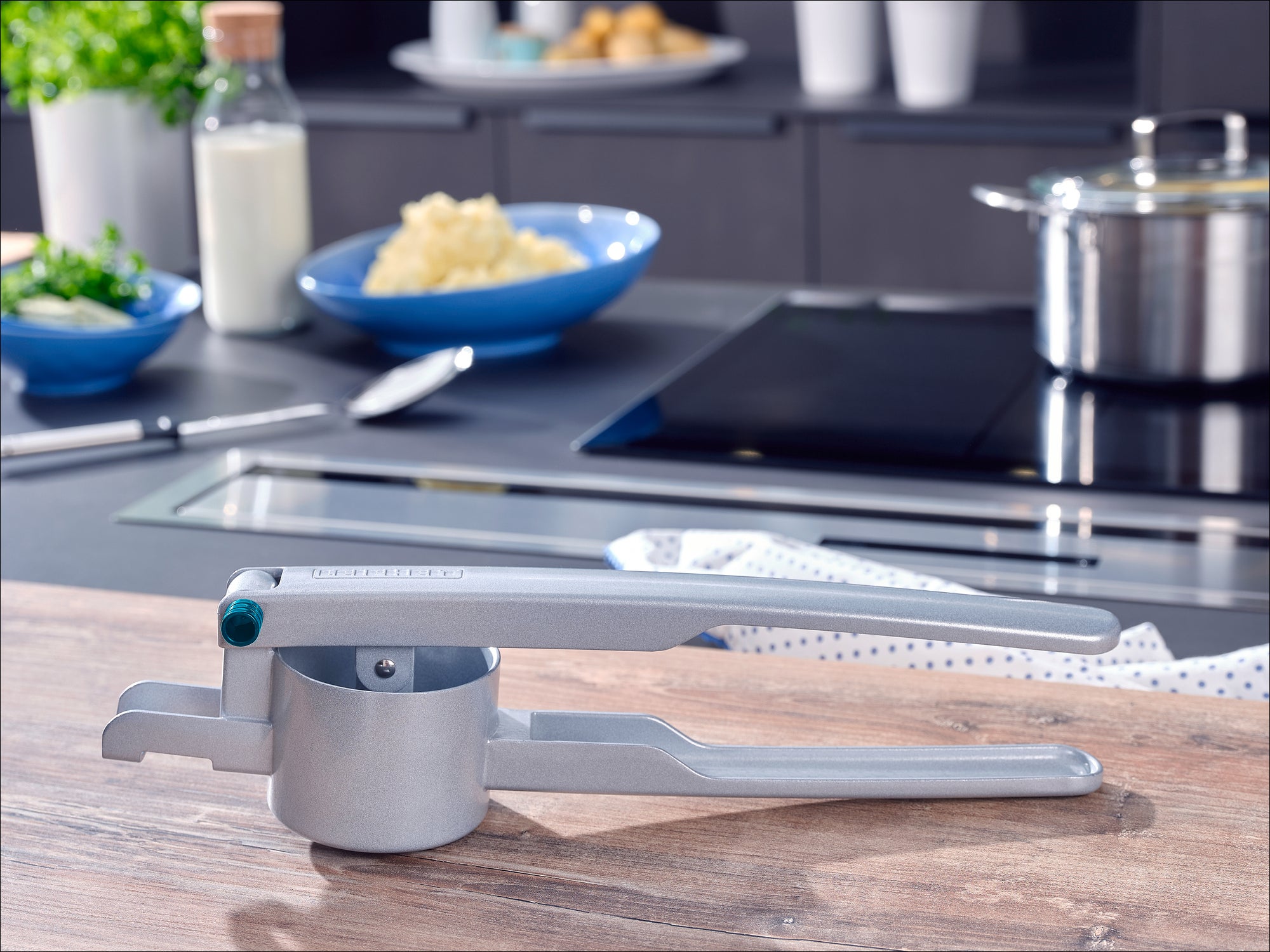 Un presse-purée en argent est posé sur un plan de travail de cuisine. À l'arrière-plan, on aperçoit une plaque de cuisson, une casserole et des bols contenant des ingrédients. Idéal pour préparer une purée de pommes de terre fraîche.