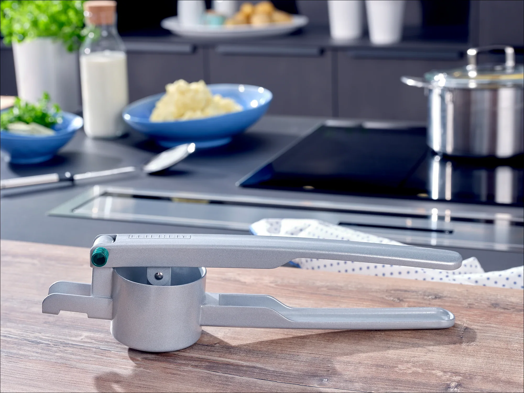 Srebrna maszynka do ziemniaków leży na blacie kuchennym. W tle widać płytę grzewczą, garnek i miski ze składnikami. Idealny do przygotowywania świeżego puree ziemniaczanego.