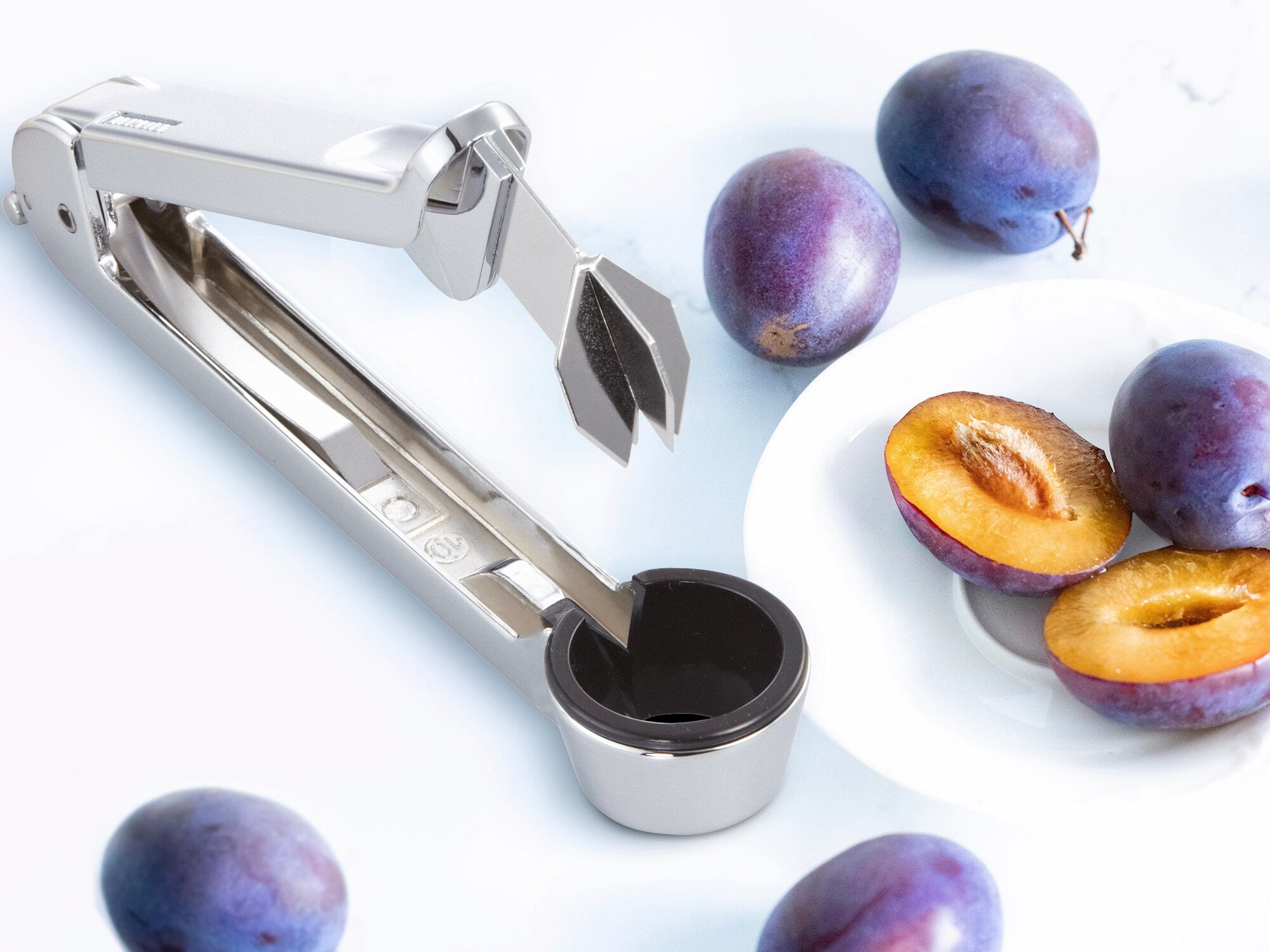 Dénoyauteur de prunes en acier inoxydable à côté d'une assiette avec des prunes coupées en deux et entières. L'appareil est idéal pour retirer rapidement et facilement les noyaux.