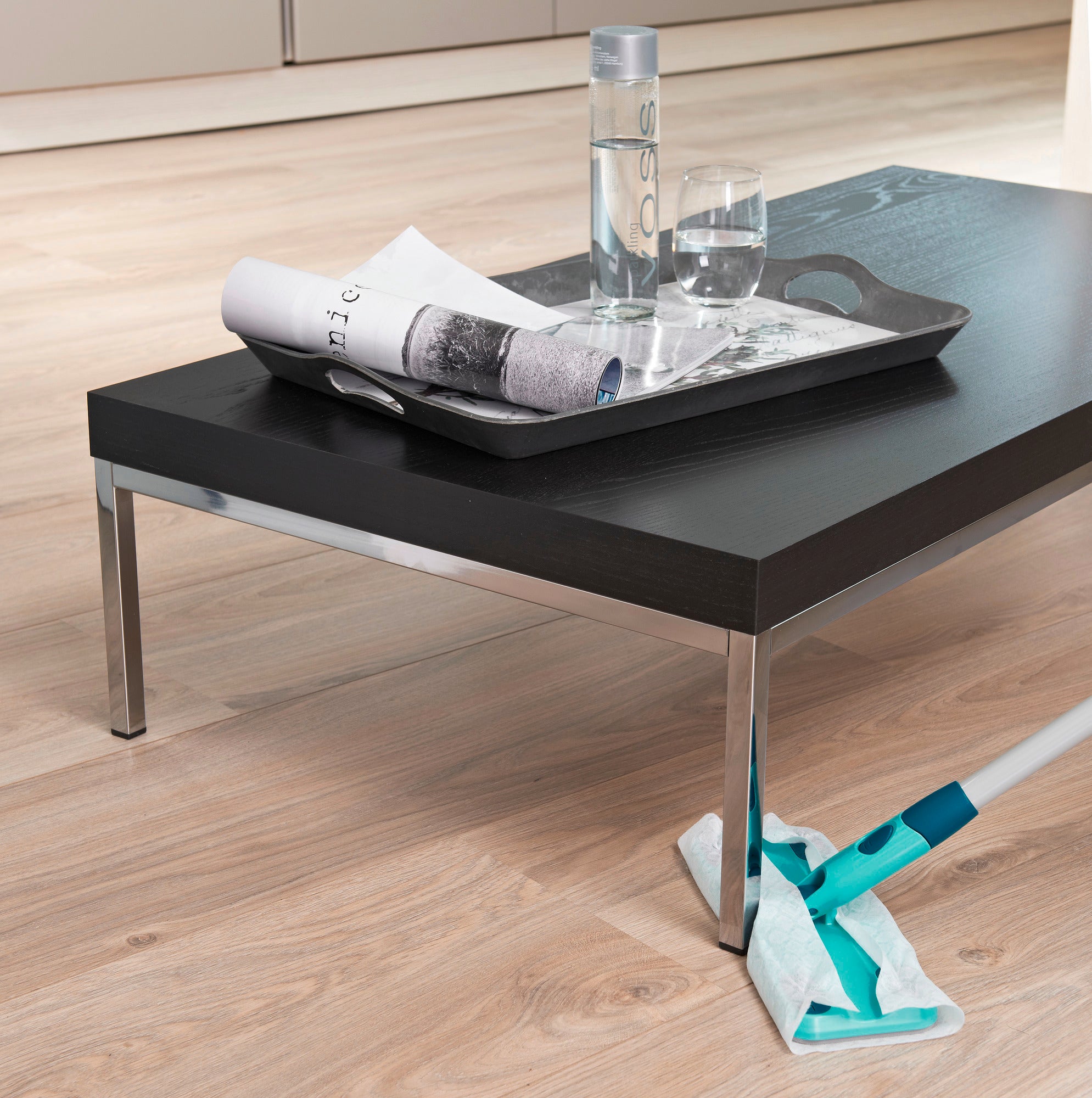 Ein moderner Wohnzimmertisch mit einem Tablett, das eine Wasserflasche und ein Glas enthält. Darunter wird der Boden mit dem Clean and Away Set gewischt. Ideal für eine saubere Umgebung.