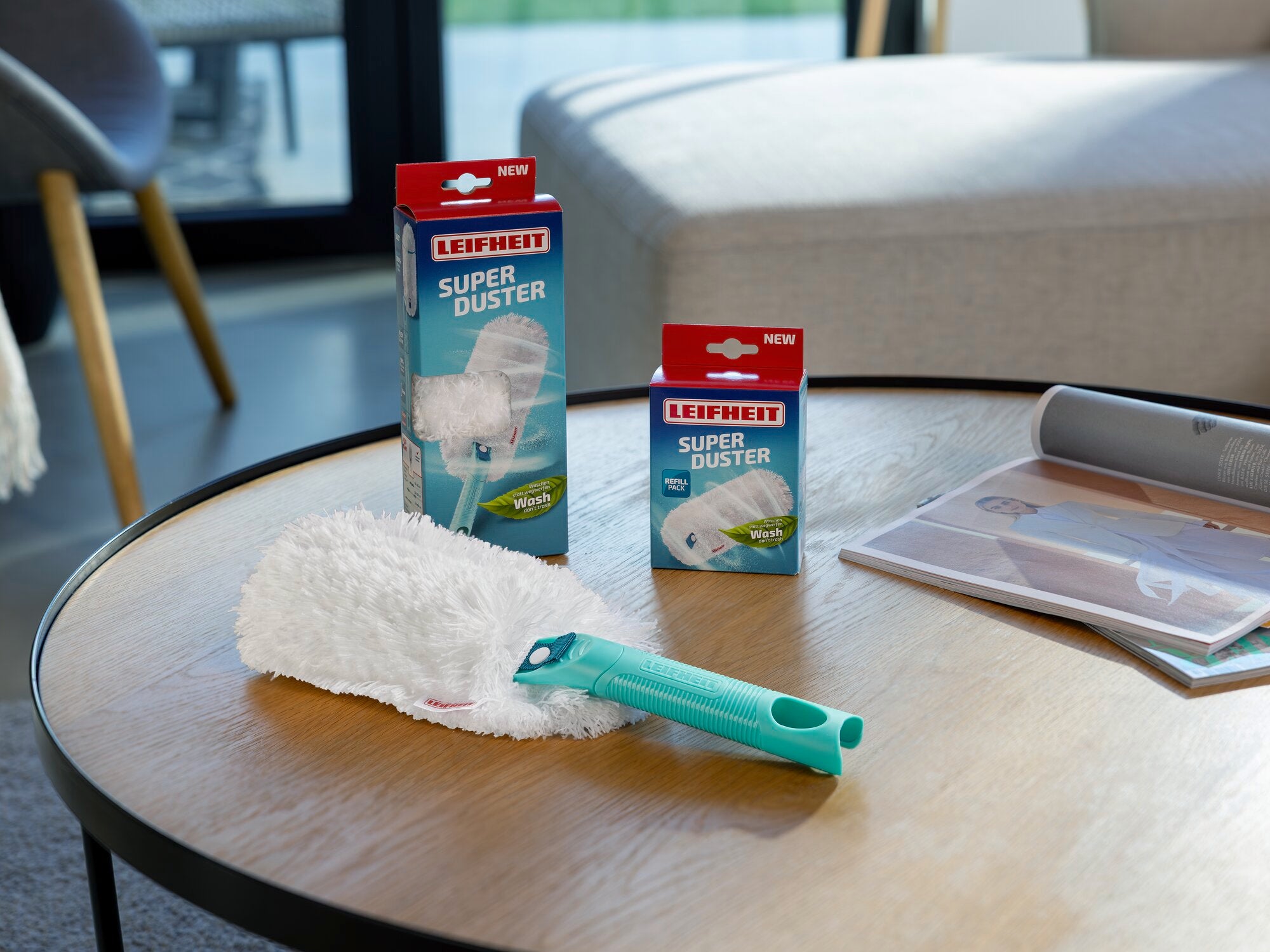 Ein SUPERDUSTER Staubwedel liegt auf einem Holztisch in einem modernen Wohnzimmer. Im Hintergrund sind zwei Verpackungen des Produkts zu sehen, die seine Anwendung und Effizienz betonen.
