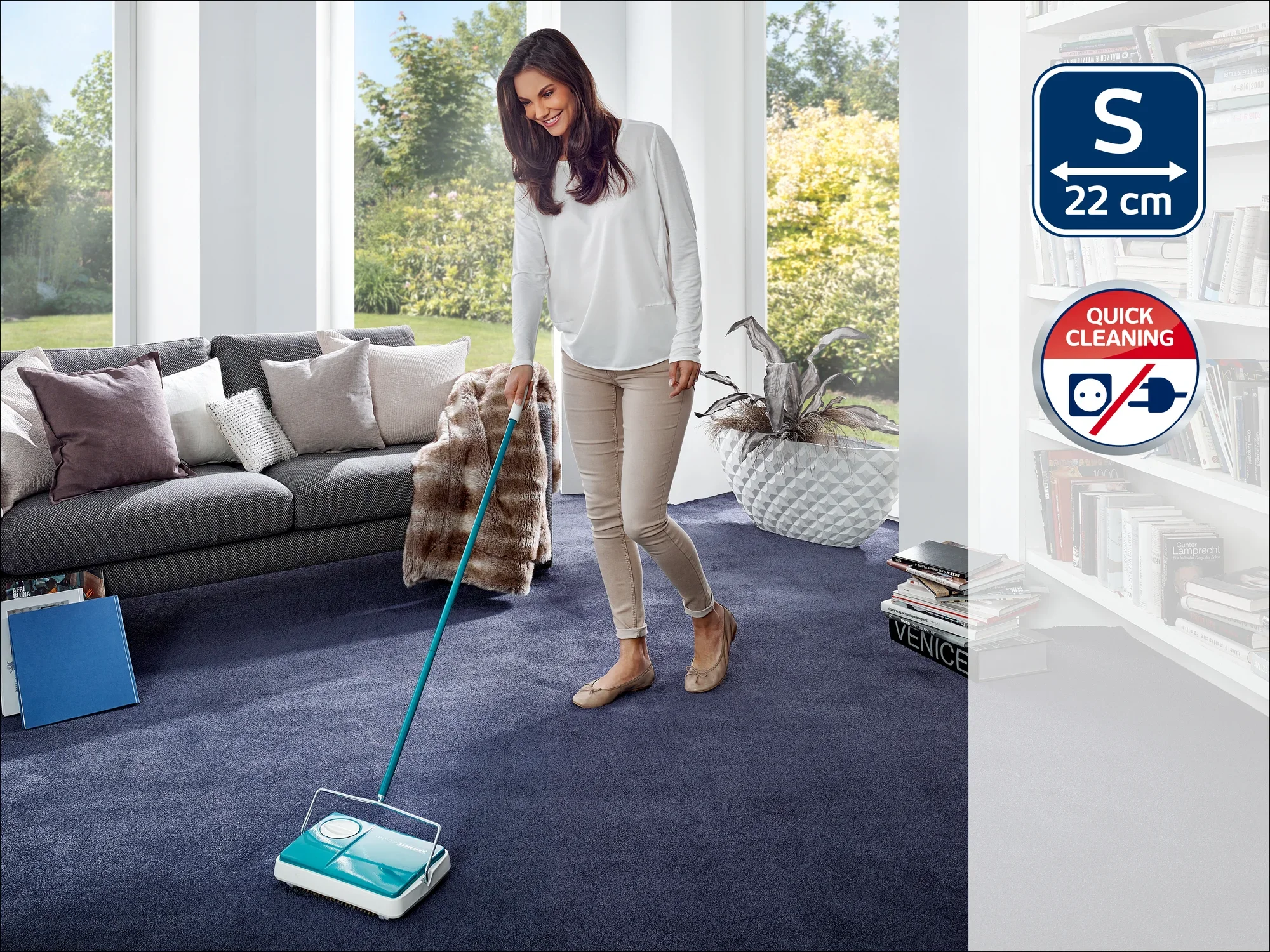 Een vrouw reinigt een donkerblauw tapijt in een moderne woonkamer met een Leifheit tapijtveger. De kamer is licht en stijlvol ingericht, met een groot raam op de achtergrond.