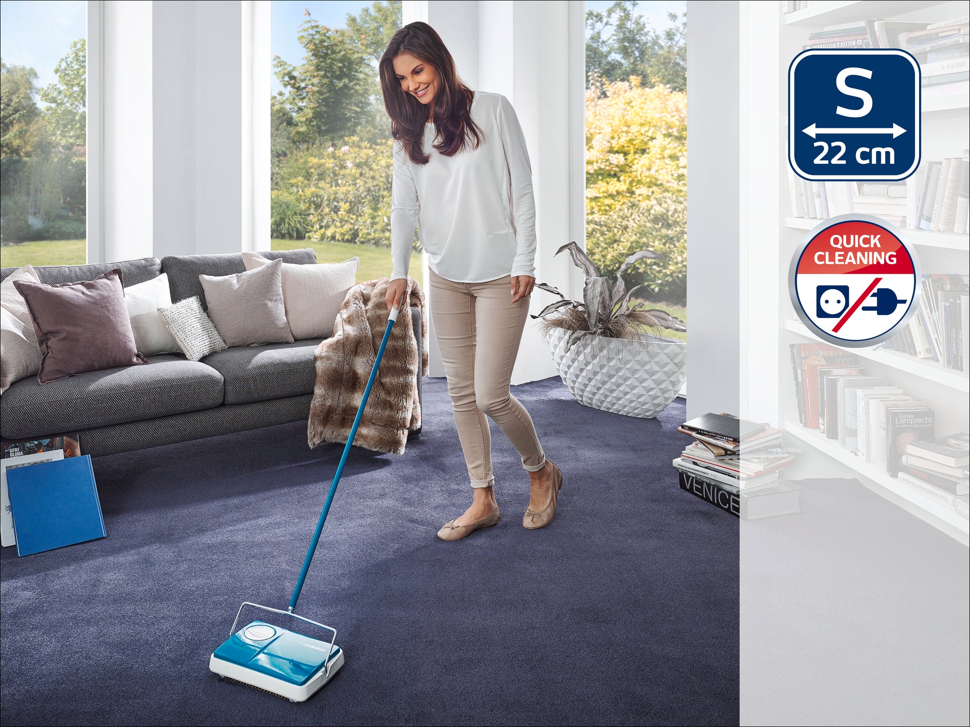 A woman is cleaning a dark blue carpet in a modern living room with a Leifheit carpet sweeper. The room is bright and stylishly furnished, with a large window in the background.