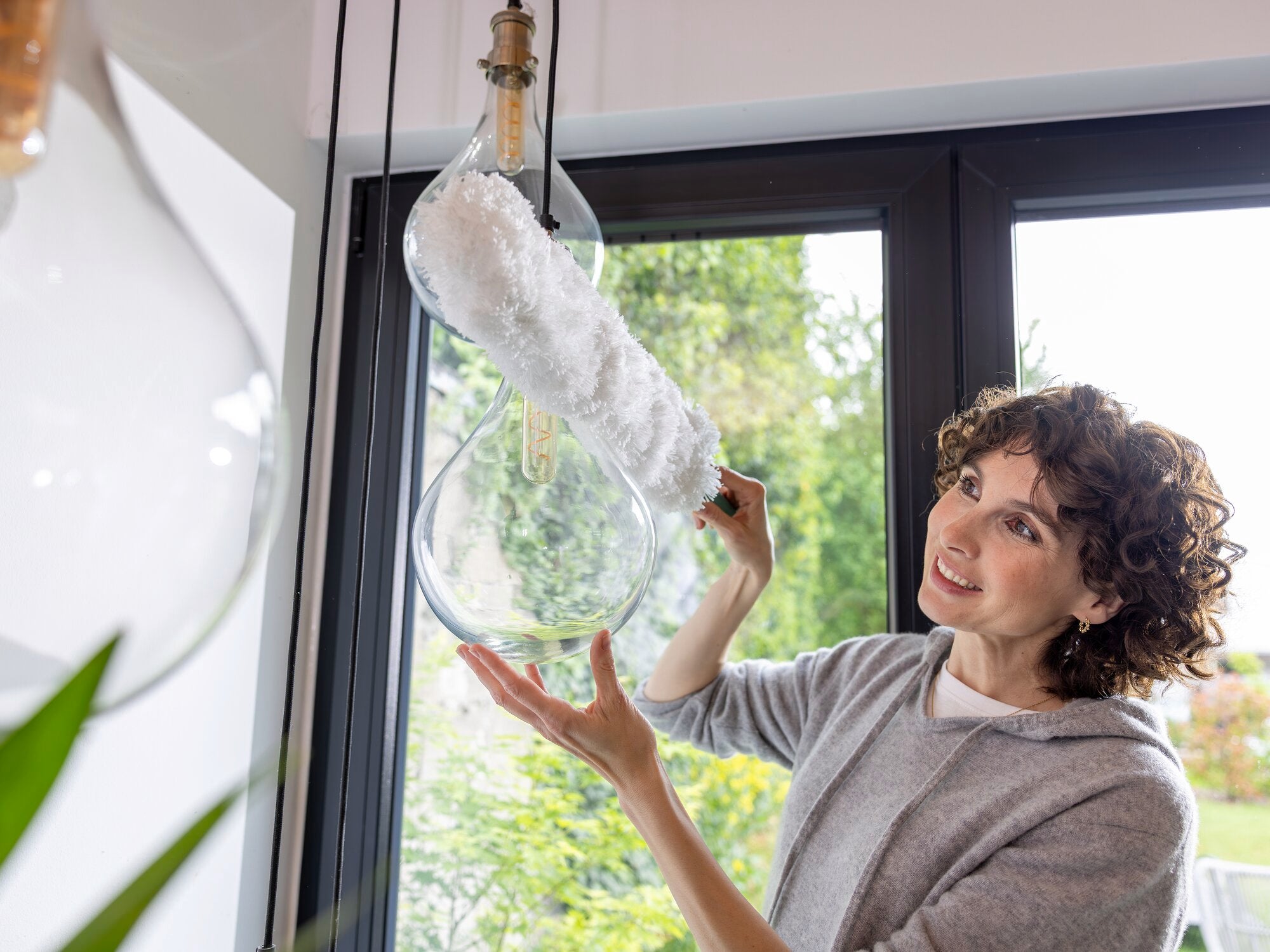 Une femme utilise le plumeau SUPERDUSTER XL pour nettoyer une grande ampoule dans une pièce moderne. Le plumeau atteint sans effort les endroits difficiles d'accès.