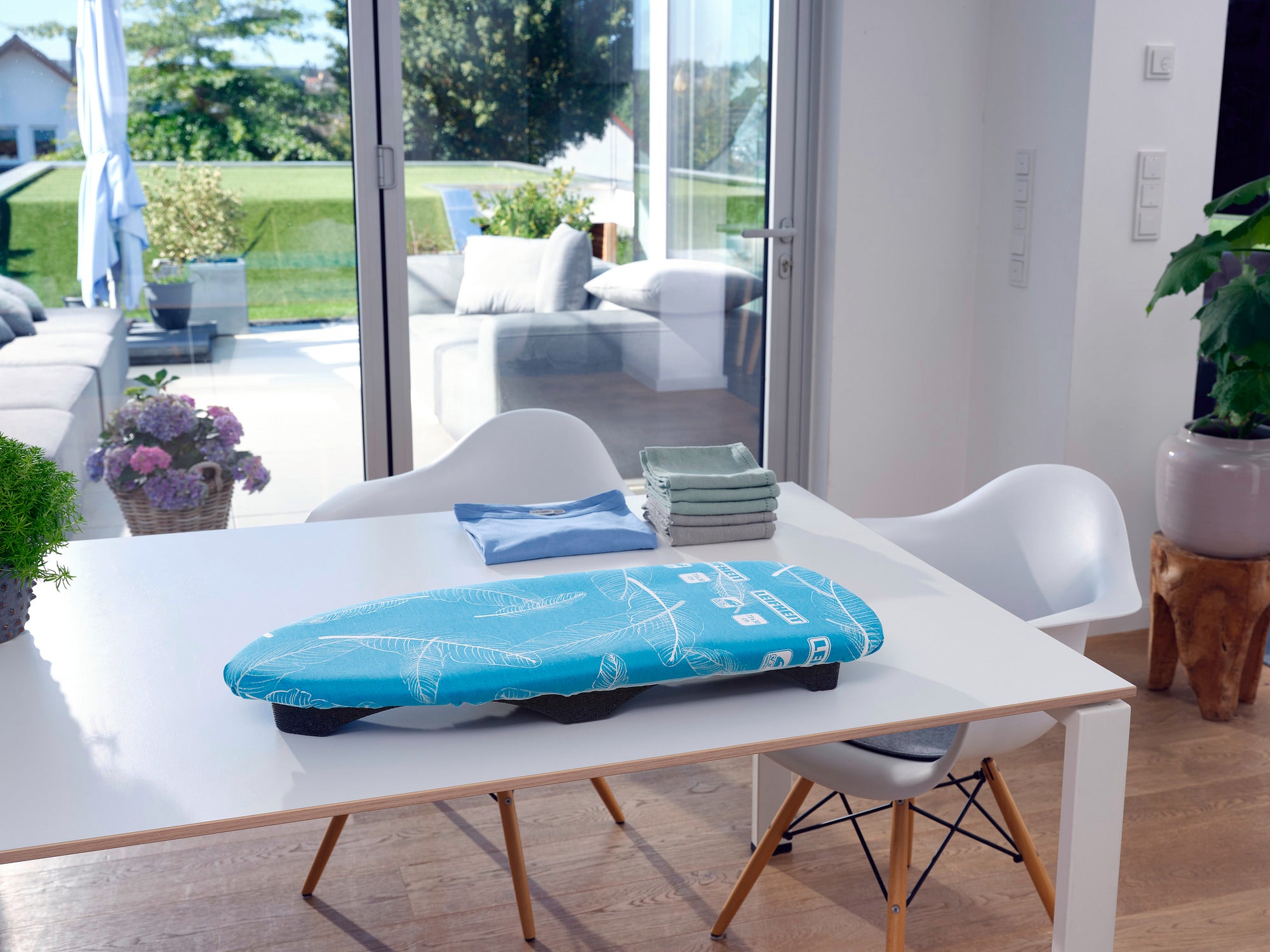 Bügeltisch Air Board Table Compact auf einem modernen weißen Tisch in einem hellen Raum mit großen Fenstern. Im Hintergrund sind ein Garten und eine Terrasse sichtbar.