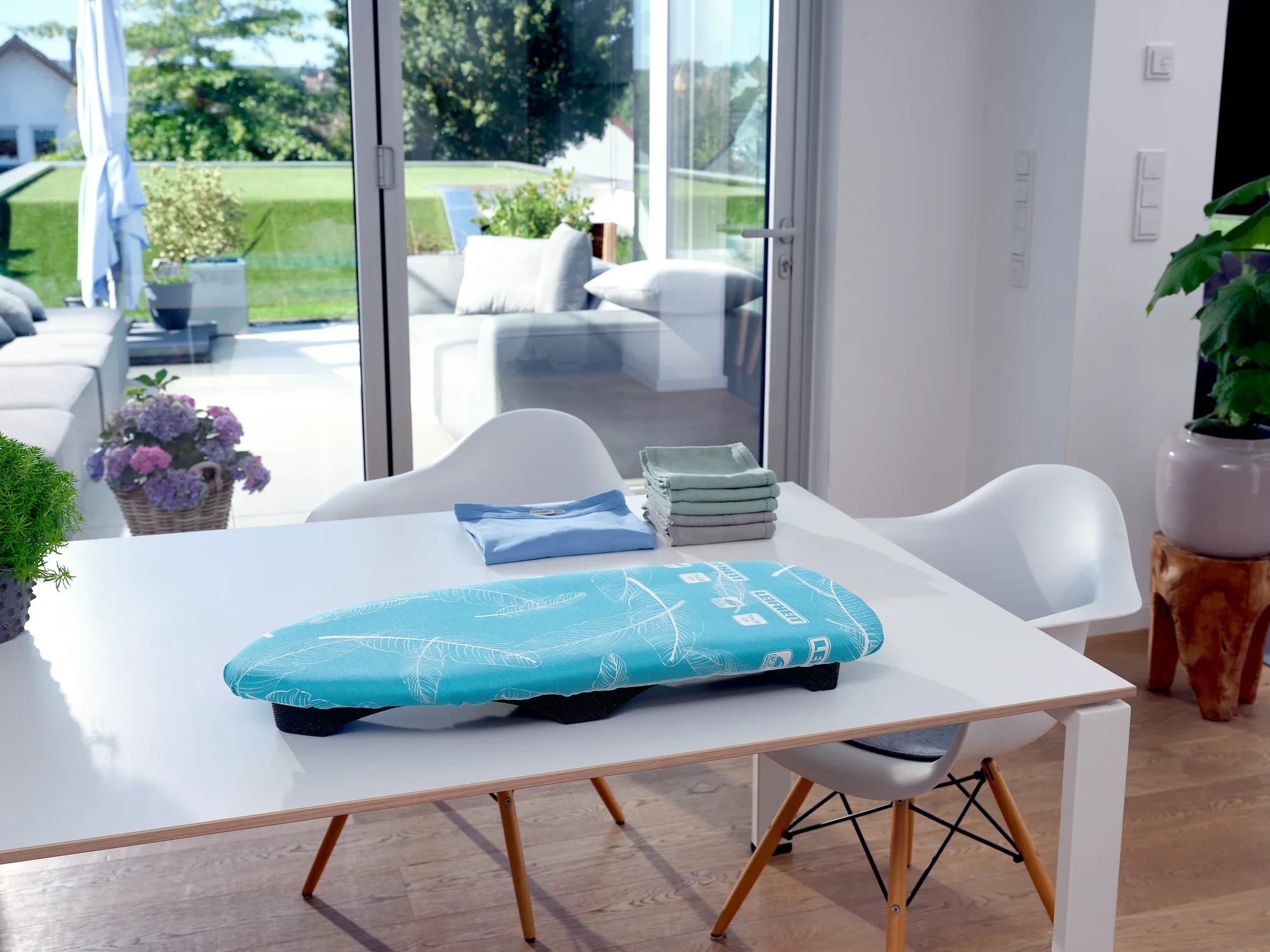 Air Board Table Compact ironing table on a modern white table in a bright room with large windows. A garden and terrace are visible in the background.